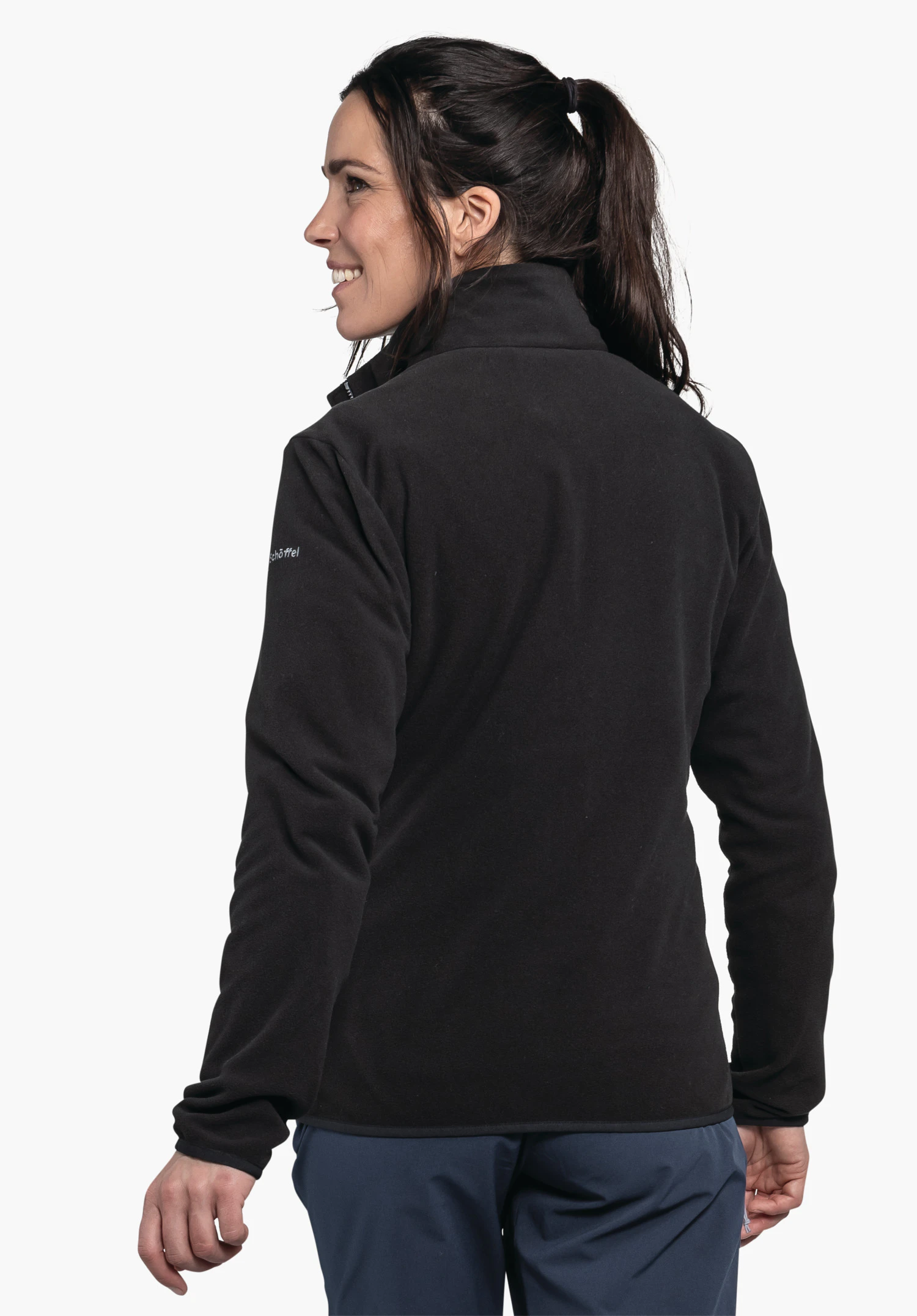 Fleece Jk Style Ash WMS zwarte fleecejas, gedragen door een vrouw in halfprofiel met blik naar links, donker haar in paardenstaart, donkerblauwe broek zichtbaar; context: lichte neutrale achtergrond. Tekst op de mouw: "Schöffel".