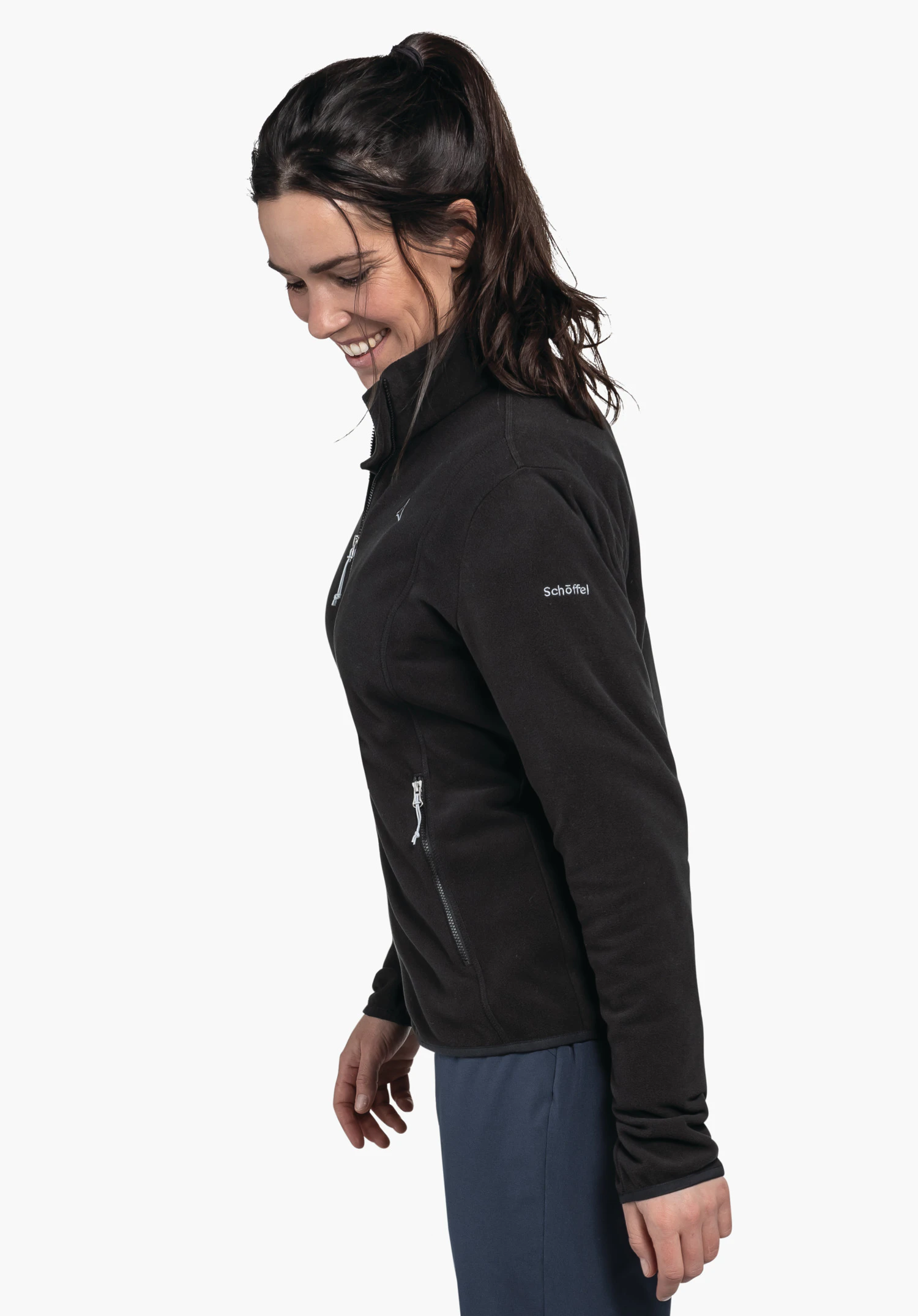 Fleece Jk Style Ash WMS zwarte fleecejas; vrouw draagt deze, glimlacht en kijkt naar beneden, staat voor een witte achtergrond; Schöffel-logo op de borst, tekst "Schöffel" op de mouw; donkerblauwe broek.
