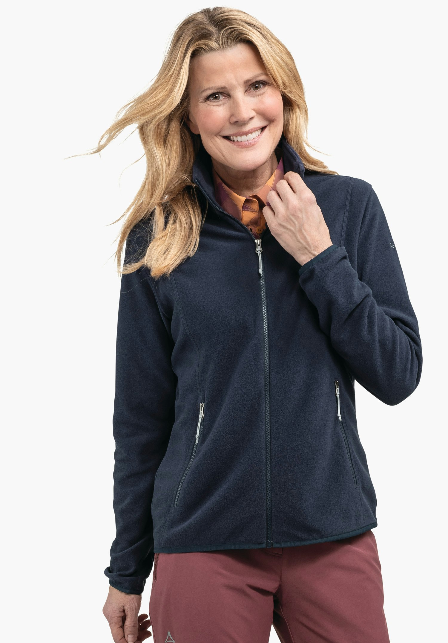 Fleece Jk Style Ash WMS Veste polaire bleu foncé portée par une femme souriante tenant le zip au col; fond clair; chemise orange visible, pantalon rouge avec logo Schöffel.