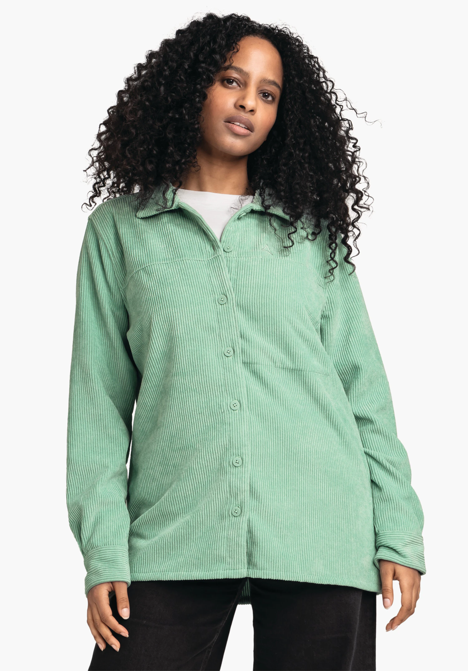 Blouse Style Lodos WMS chemise en velours côtelé vert clair, boutonnée à l’avant et portée ample par une femme; logo Schöffel sur la poitrine, t-shirt blanc, pantalon noir, longs cheveux noirs bouclés; fond blanc.