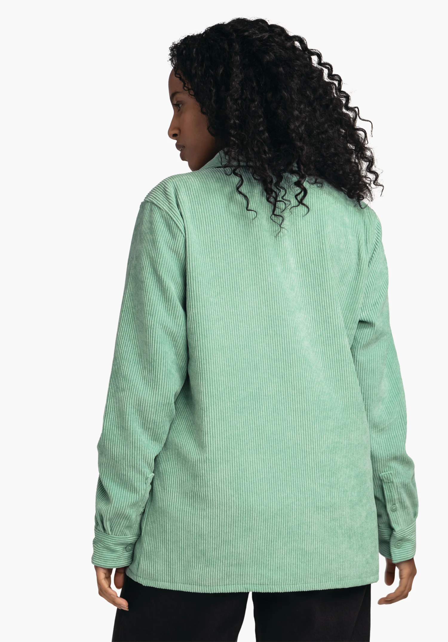 Blouse Style Lodos WMS lichtgroene corduroy overhemdjas; gedragen door een staande persoon in halfprofiel met zichtbare rug, donker krullend haar valt over de kraag, zwarte broekrand, neutrale lichte achtergrond.