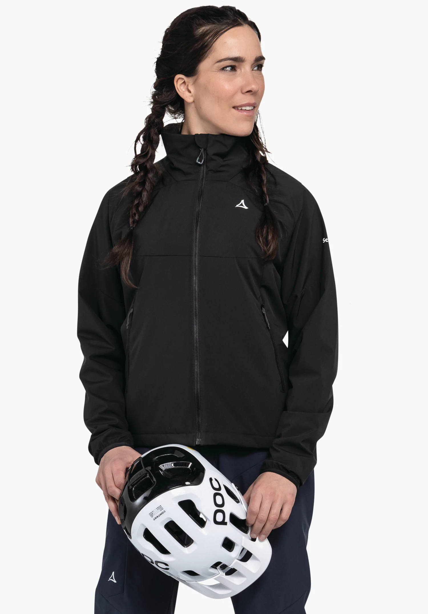 Ins Jacket Style Salza WMS personne portant une veste noire avec logo Schöffel, tenant un casque blanc avec inscription POC à deux mains; debout devant un fond blanc, regard vers la droite, cheveux tressés.