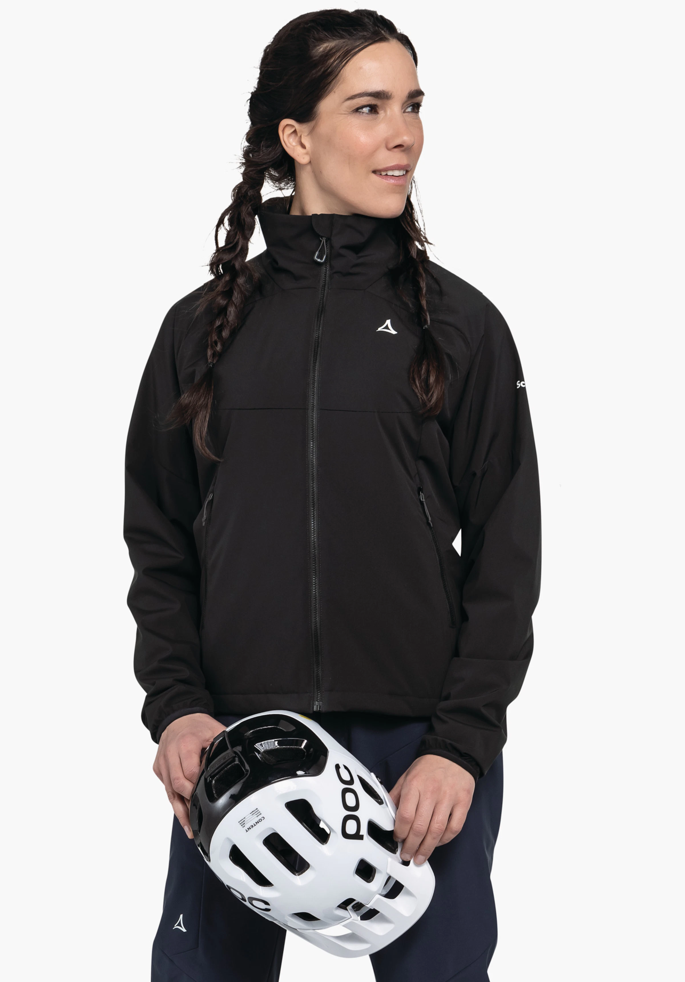 Ins Jacket Style Salza WMS personne portant une veste noire avec logo Schöffel, tenant un casque blanc avec inscription POC à deux mains; debout devant un fond blanc, regard vers la droite, cheveux tressés.