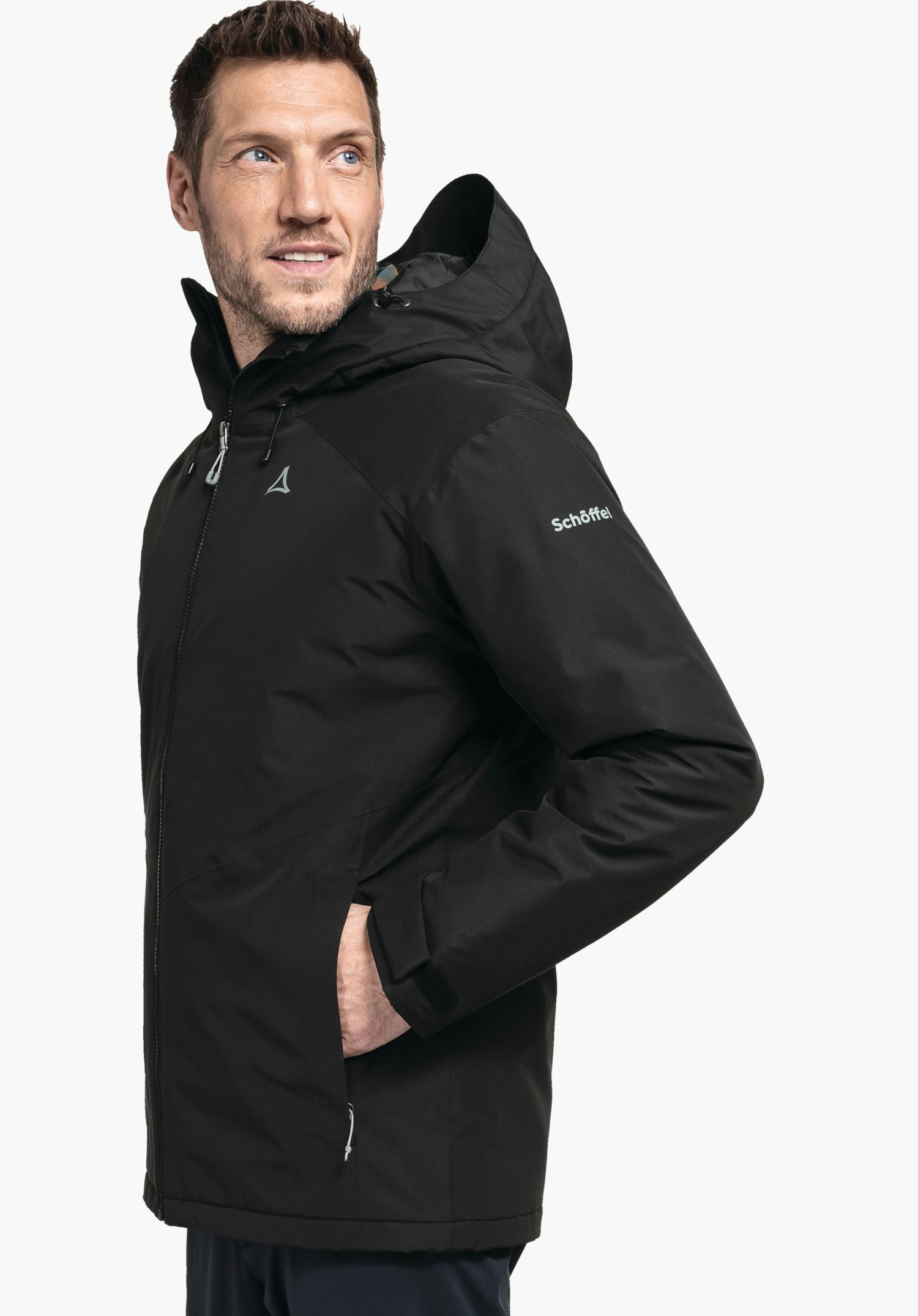 Ins Jacket Style Wildkar MNS Schwarze Kapuzenjacke mit Schöffel-Logo auf Brust und Schriftzug Schöffel auf dem Ärmel, von einem Mann getragen, Hand in Tasche, stehender Blick, weißer Hintergrund.