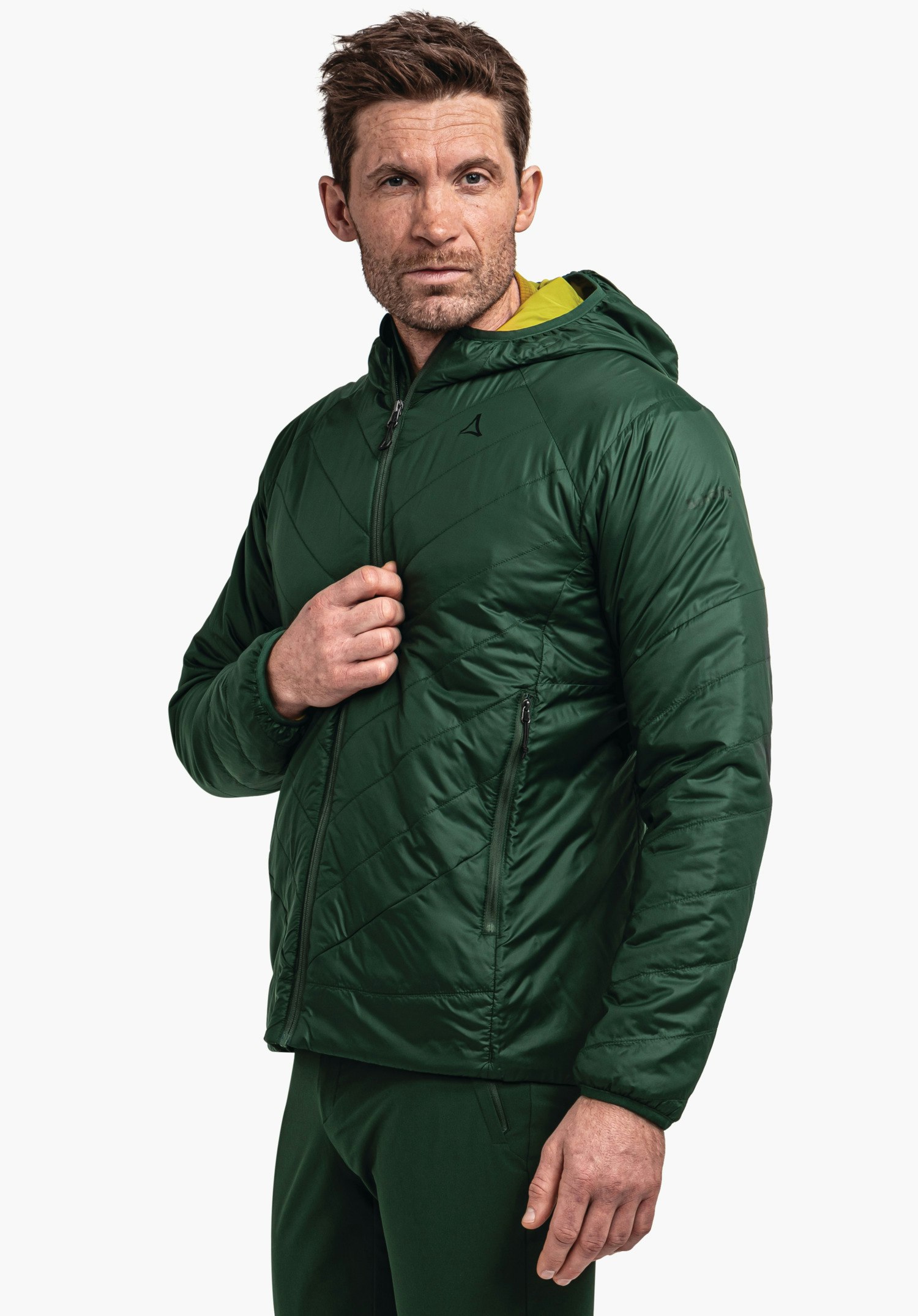 Ins Jacket Style Cascata MNS dunkelgrüne gesteppte Kapuzenjacke (Schöffel-Logo) wird von Mann halbseitig getragen, eine Hand am Reißverschluss, hellgelbes Innenfutter sichtbar; neutraler weißer Hintergrund.