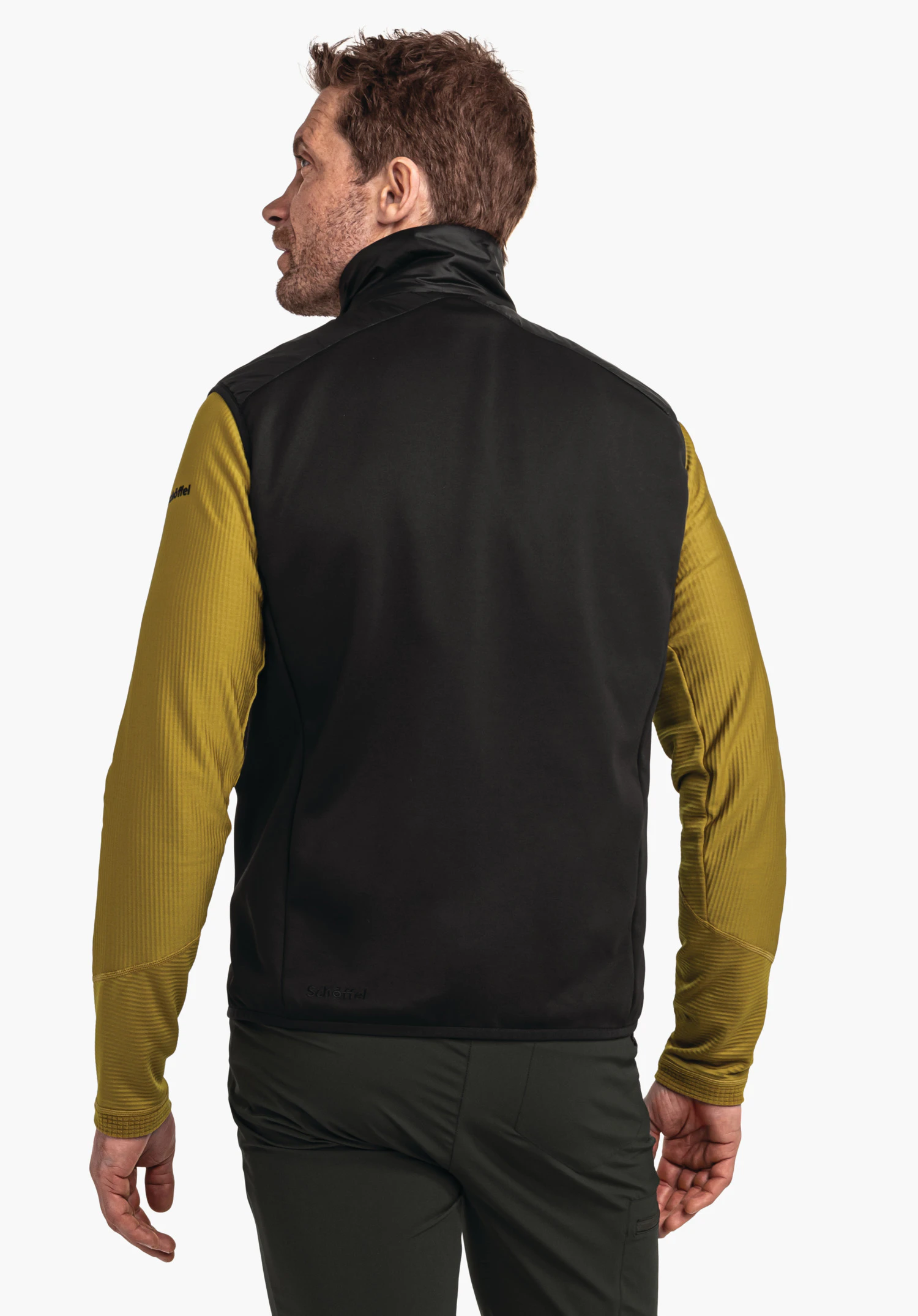 Hybrid Vt Style Cascata MNS black vest worn over a dark yellow long-sleeve top; man stands looking over his left shoulder; white background. Vest text: “Schöffel”., Hybrid Vt Style Cascata MNS black vest worn over a dark yellow long-sleeve top; man stands looking over his left shoulder; white background. Vest text: “Schöffel”.