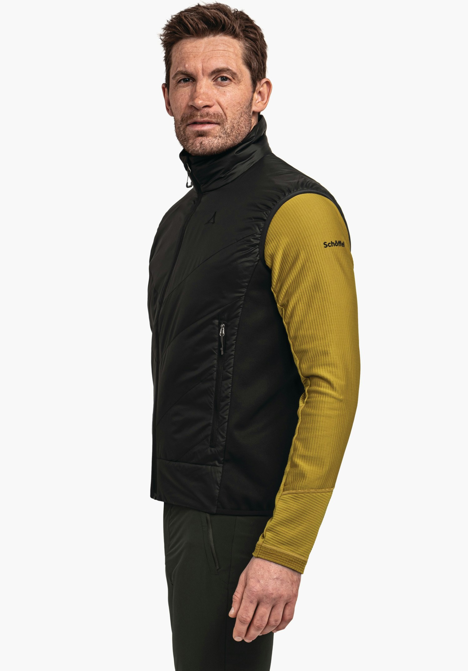 Hybrid Vt Style Cascata MNS gilet noir avec logo Schöffel sur la poitrine porté par un homme debout de profil, regard vers la caméra; sous-couche manches longues jaune, pantalon vert foncé, fond blanc. Texte: "Schöffel".