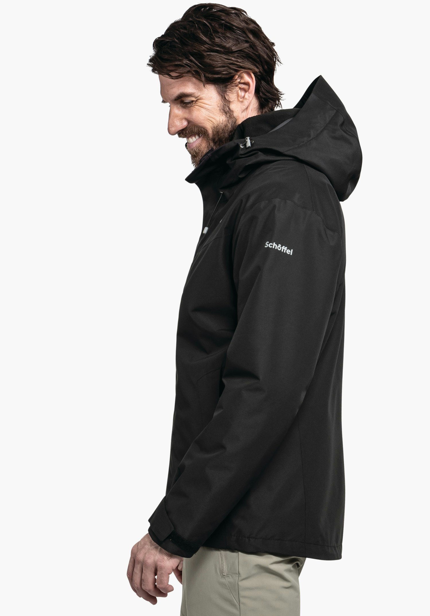 3in1 Jacket Style Okere MNS schwarze Kapuzenjacke, getragen von einem lächelnden Mann im Profil mit leicht geneigtem Kopf, Hände entspannt; hellweißer Hintergrund; Schriftzug "Schöffel" auf dem Ärmel.