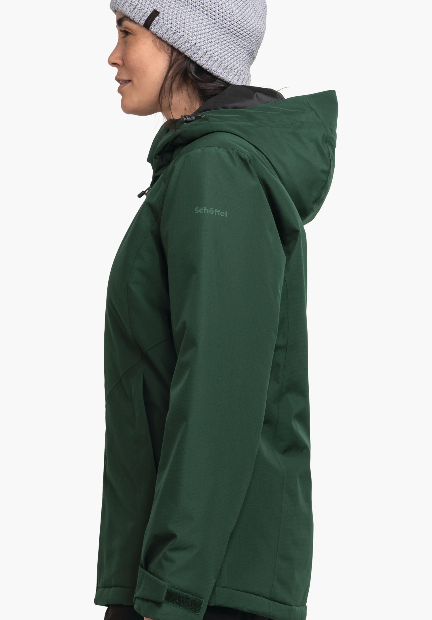 Ins Jacket Style Wildkar WMS veste vert foncé portée par une personne debout de profil regardant vers la gauche; bonnet tricoté gris clair; fond blanc neutre. Texte: "Schöffel".