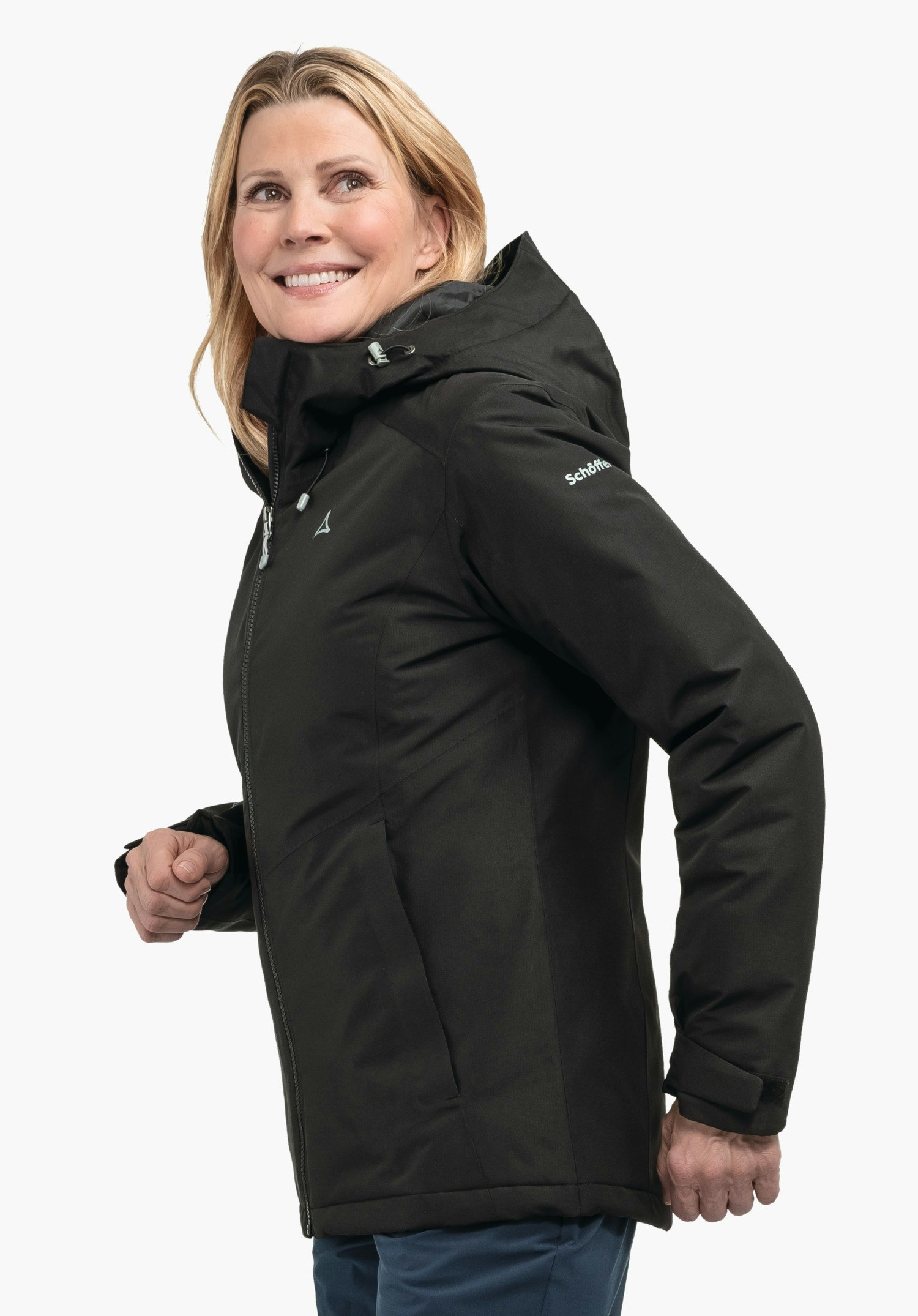 Ins Jacket Style Wildkar WMS veste noire à capuche (logo Schöffel sur la poitrine, inscription "Schöffel" sur la manche) portée par une femme souriante en mouvement vers l’avant; fond blanc.