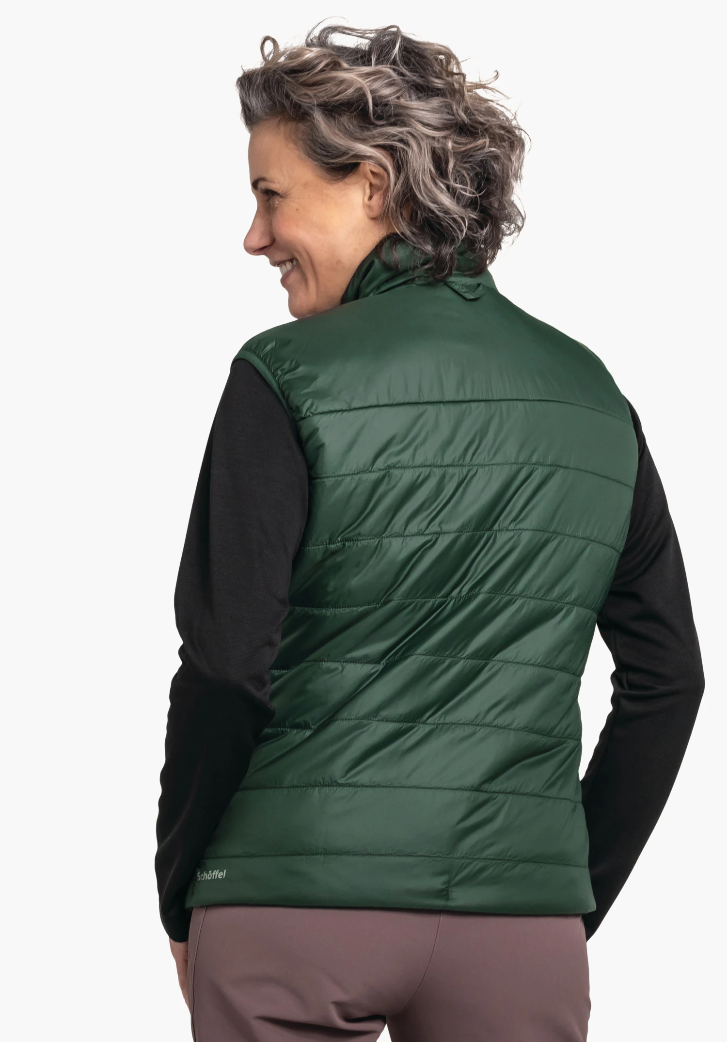 Ins Vest Style Yew WMS dark green quilted vest worn by a smiling woman in half profile; black base layer, purple trousers; white background. Text: “Schöffel”.