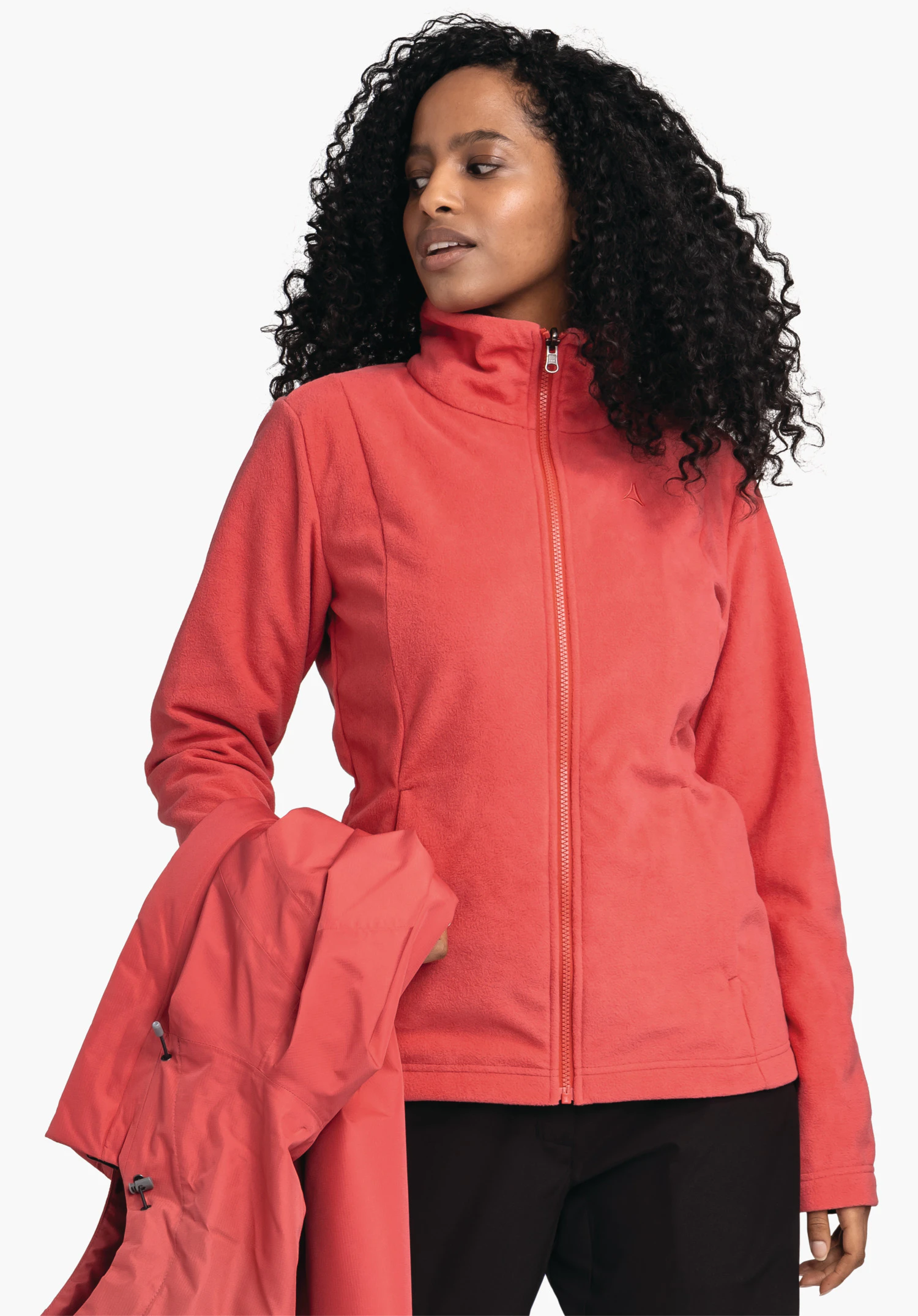 3in1 Jacket Style Okere WMS hellrote Fleecejacke mit Schöffel-Logo; Frau trägt sie, hält eine hellrote Jacke in der linken Hand, steht vor weißem Hintergrund und trägt schwarze Hosen.
