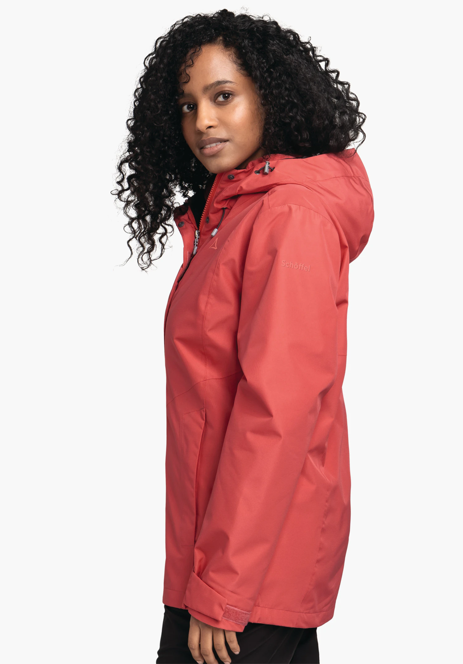 3in1 Jacket Style Okere WMS rote Jacke, seitlich getragen von einer Frau mit schwarzem, lockigem Haar, Kapuze aufgesetzt, vor hellem Hintergrund; Schöffel-Logo auf der Brust; Schriftzug auf dem Ärmel: "Schöffel".