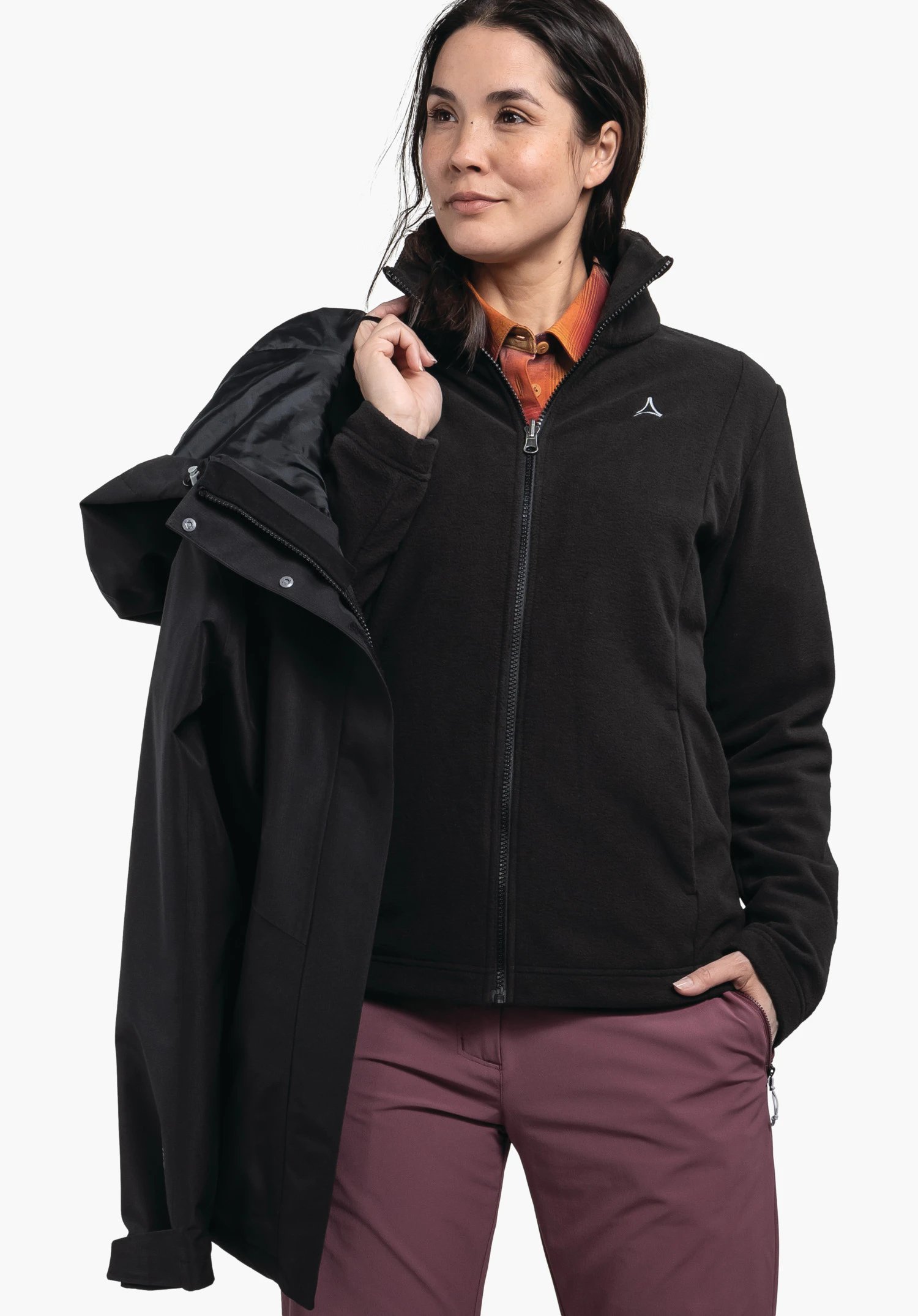 3in1 Jacket Style Okere WMS Frau trägt schwarze Fleecejacke mit Schöffel-Logo, hält schwarze Regenjacke über der Schulter, schaut nach rechts; darunter dunkel orange Hemd und dunkel lila Hose, weißer Hintergrund.