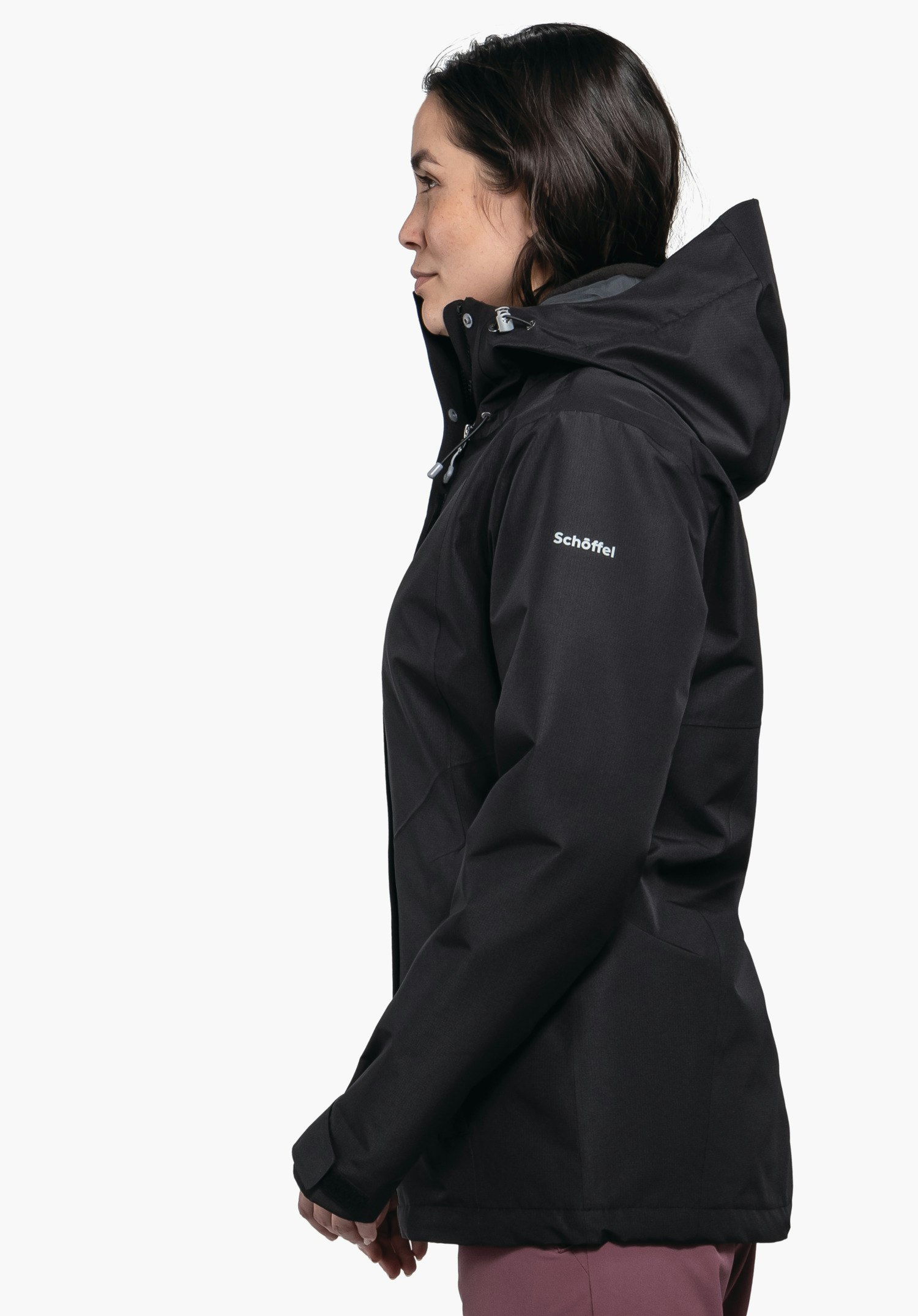 3in1 Jacket Style Okere WMS veste noire portée par une femme; elle se tient de profil et regarde vers la gauche, capuche relevée, bras détendus, fond blanc. Texte sur la manche: "Schöffel".