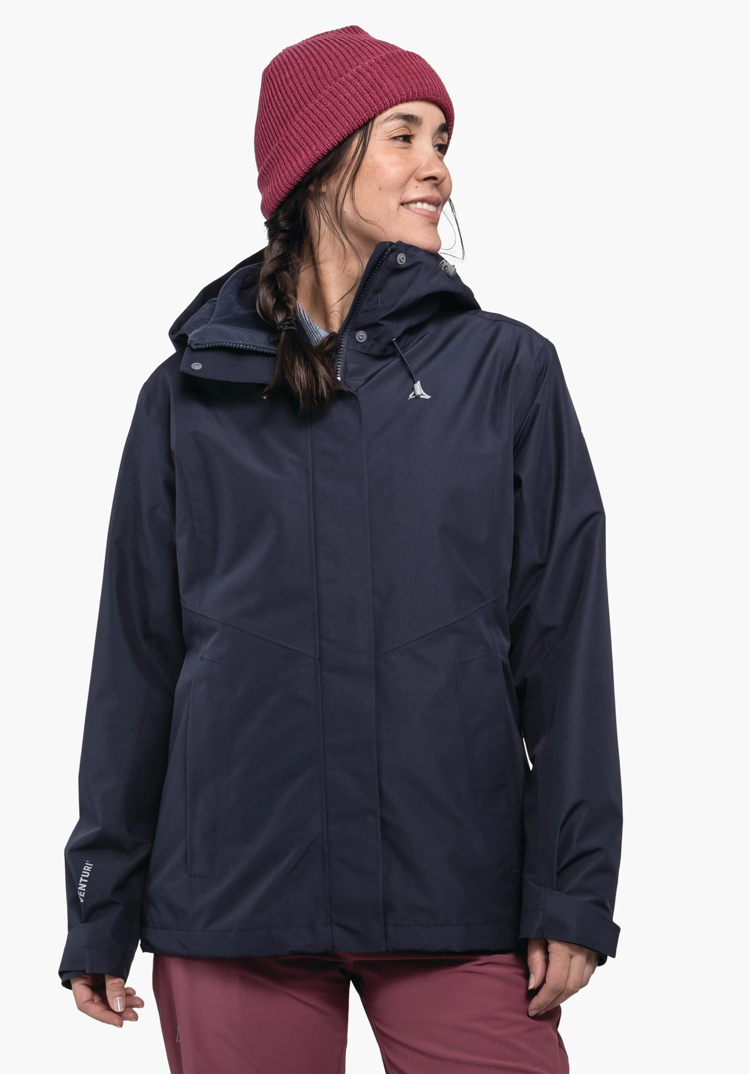 3in1 Jacket Style Okere WMS Dunkelblaue Jacke mit Schöffel-Logo, getragen von einer Frau, die nach rechts lächelt; sie trägt eine dunkelrote Mütze und rote Hosen; heller Hintergrund. Text: VENTURI