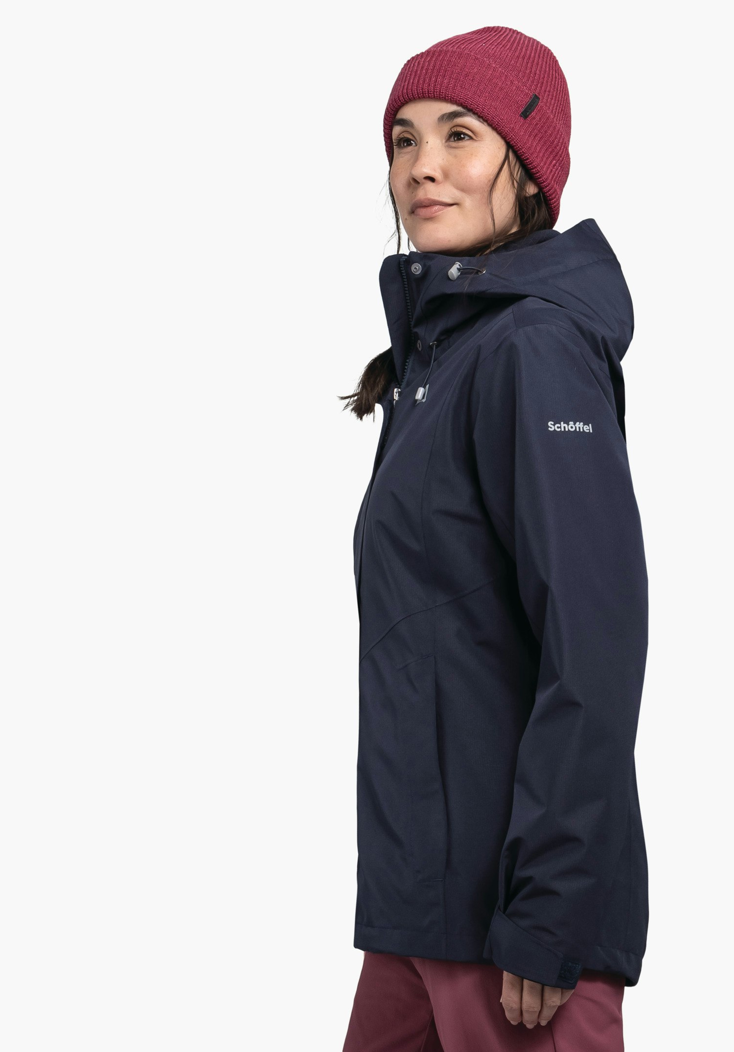 3in1 Jacket Style Okere WMS veste bleu foncé portée par une femme debout de profil, regard légèrement vers le haut; elle porte un bonnet rouge et un pantalon rouge devant fond blanc. Texte sur la manche: Schöffel.