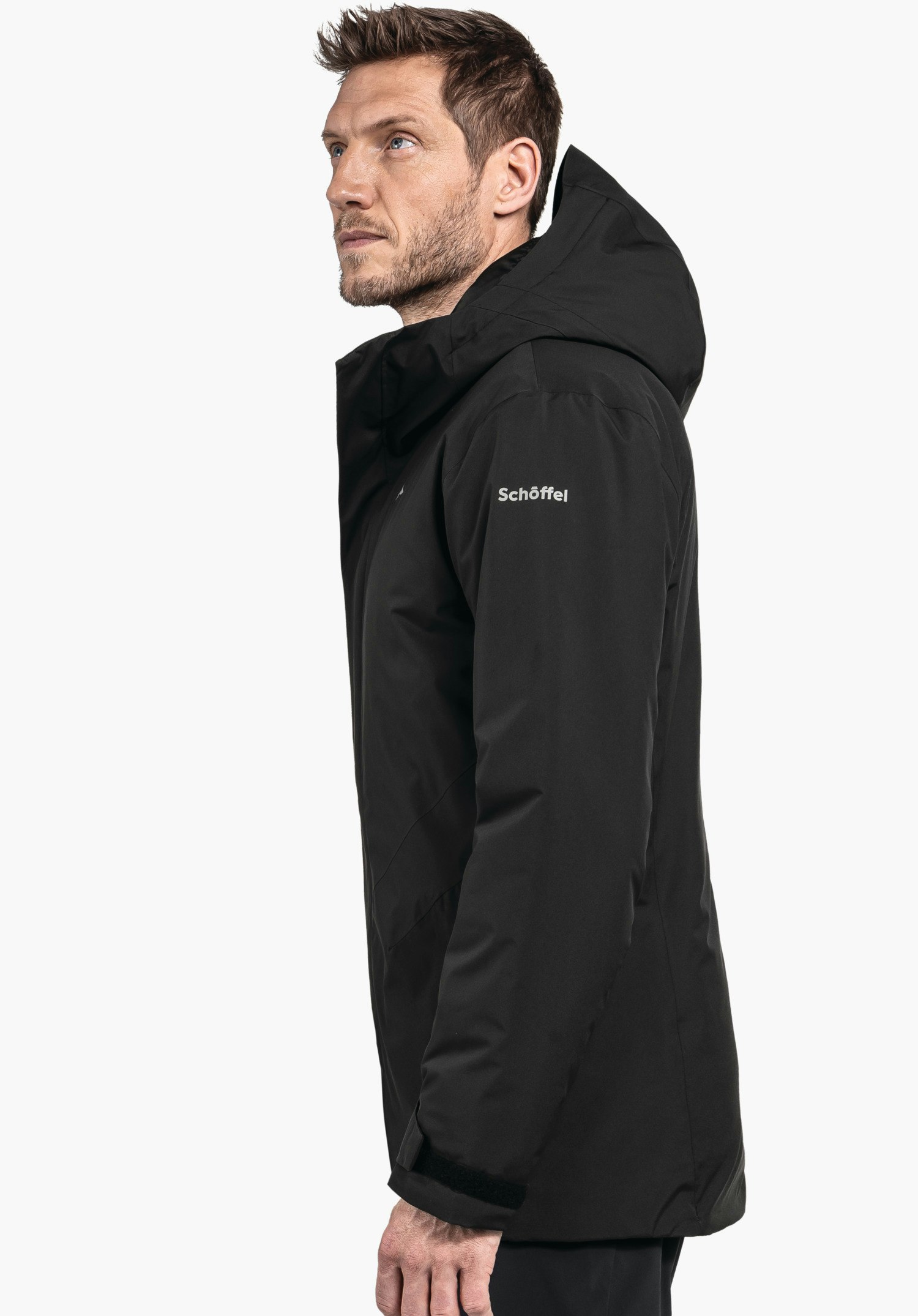 Parka Style Froda MNS Schwarze Herrenjacke mit Kapuze, getragen von einem seitlich stehenden Mann, der nach oben blickt; neutraler weißer Hintergrund. Text auf Ärmel: "Schöffel".