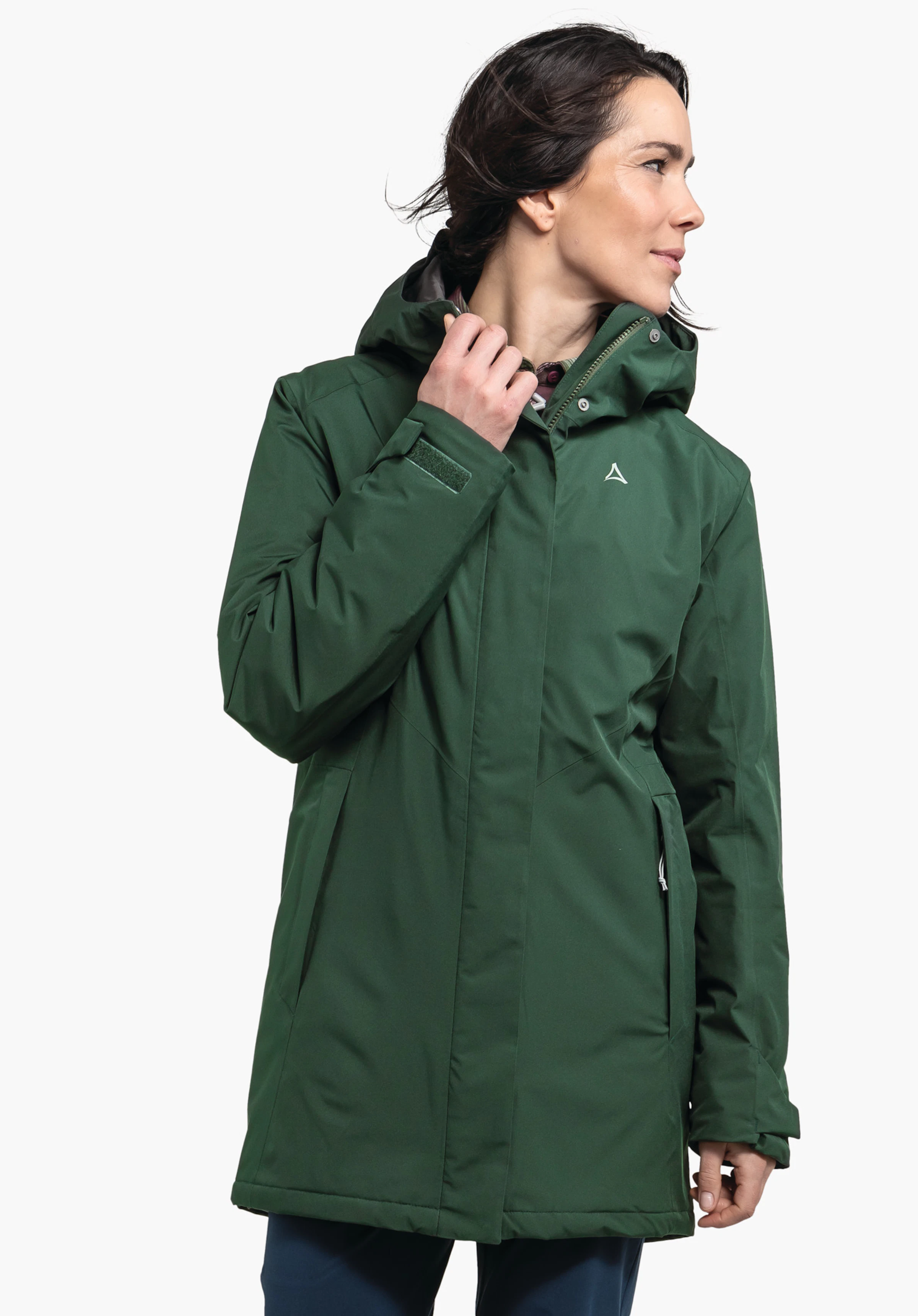 Parka Style Froda WMS dunkelgrüne Parkajacke mit Schöffel-Logo, Person zieht den Kragen hoch, Blick nach rechts, Hände am Reißverschluss, stehende Pose vor weißem Hintergrund.