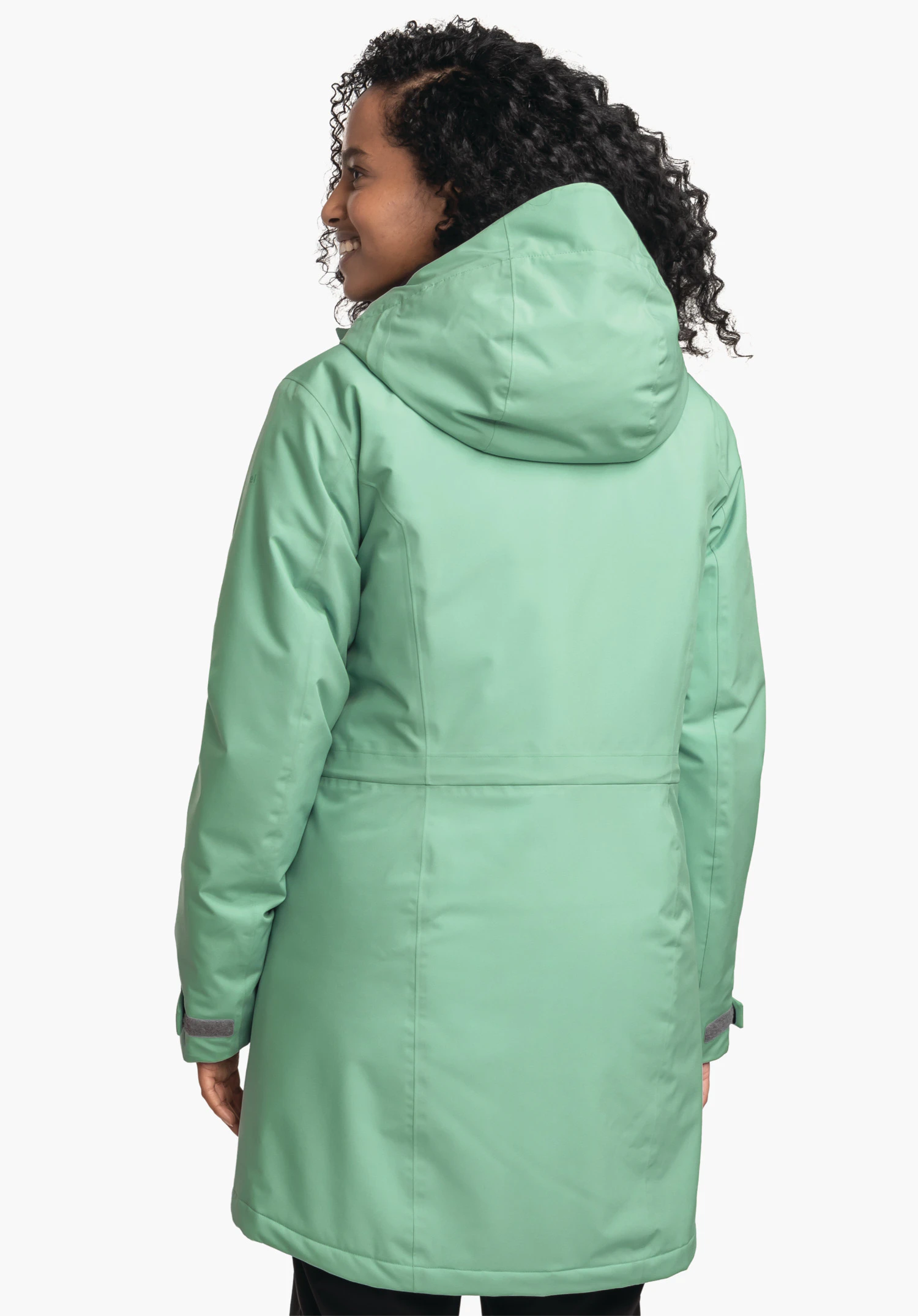 Parka Style Froda WMS parka vert clair, porté par une femme se tournant légèrement vers l’arrière; elle sourit devant un fond clair neutre; capuche reposant sur le dos, manches avec fermeture velcro.