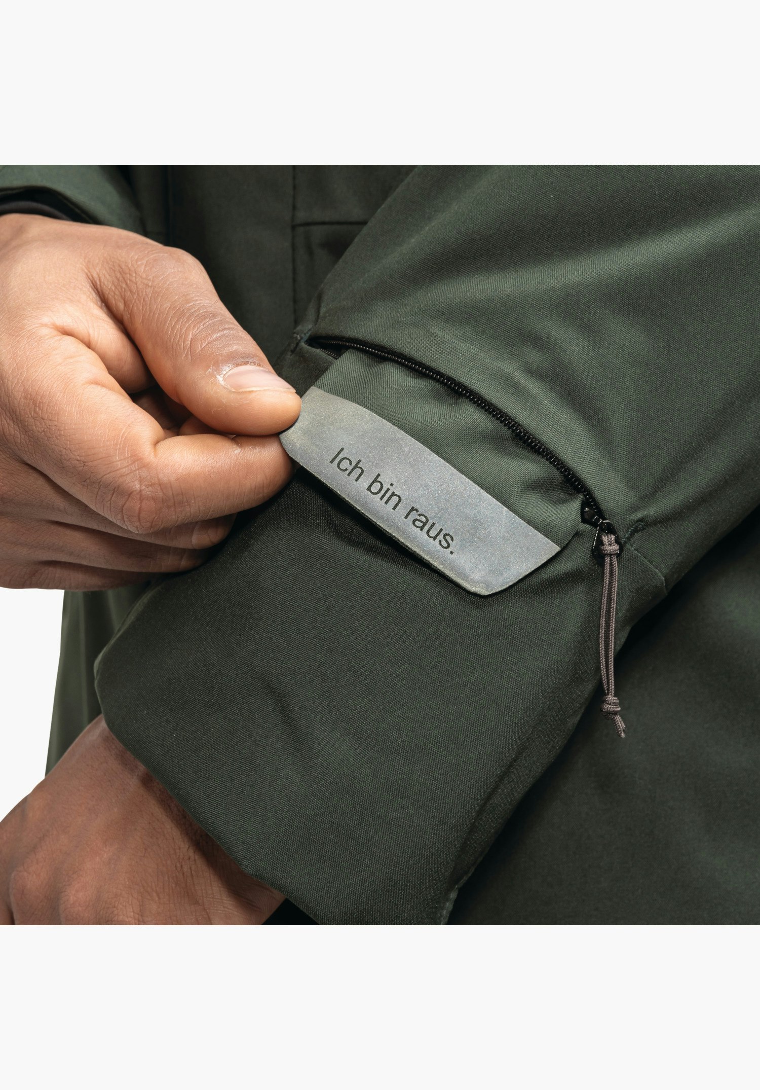Ins Parka Style Crivat MNS, Detailansicht des Ärmels mit einer kleinen Reißverschlusstasche und einem herausziehbaren Anhänger mit dem Schriftzug „Ich bin raus.“ auf grünem Material, Nahaufnahme vor weißem/neutralem Hintergrund.