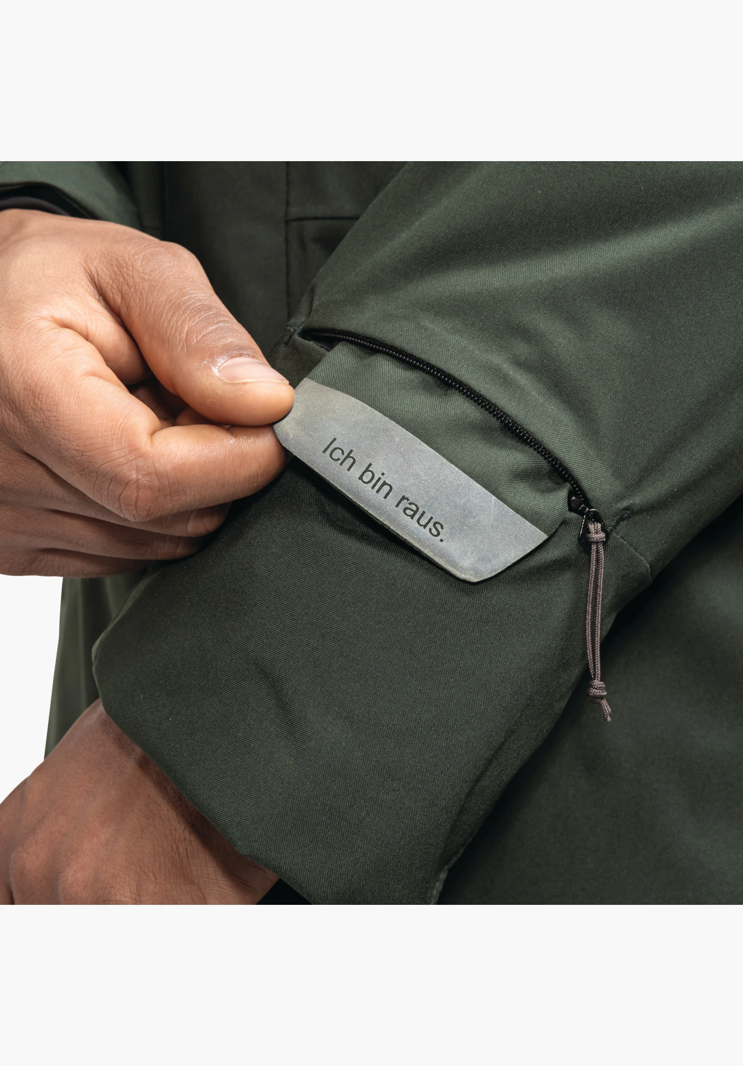 Ins Parka Style Crivat MNS, Detailansicht des Ärmels mit einer kleinen Reißverschlusstasche und einem herausziehbaren Anhänger mit dem Schriftzug â€žIch bin raus.â€œ auf grünem Material, Nahaufnahme vor weißem/neutralem Hintergrund.