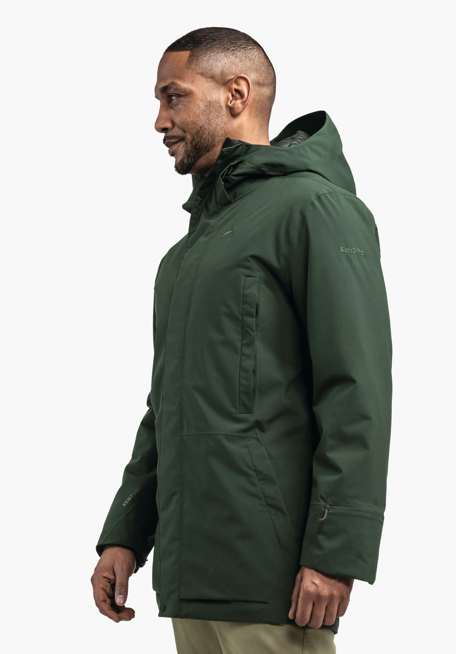 Ins Parka Style Crivat MNS Dunkelgrüne Parka (Schöffel-Logo) wird von einem seitlich stehenden Mann getragen; Kapuze aufgerichtet, Hände entspannt; neutraler heller Hintergrund. Text auf Jacke: "Schöffel", "VENTURI".