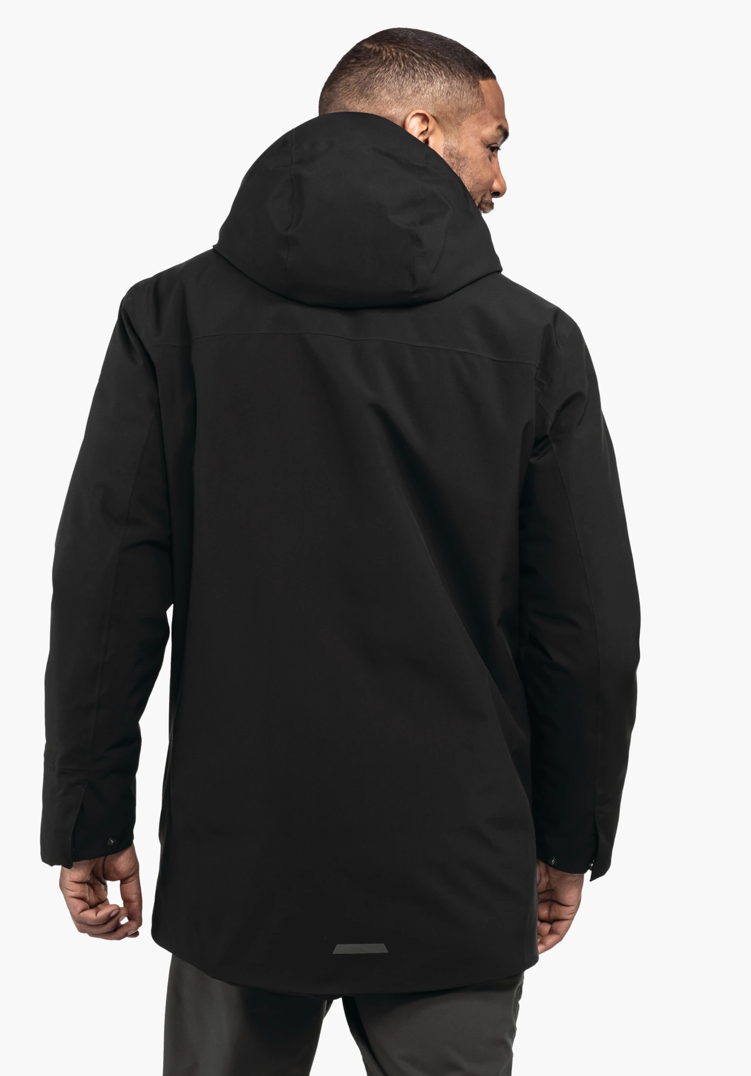 Ins Parka Style Crivat MNS Schwarze lange Kapuzenjacke, getragen von einem Mann, Rückenansicht; die Jacke hängt glatt und leicht ausgestellt. Heller weißer Hintergrund, dunkelgraue Hose, kleiner reflektierender Streifen am Saum.