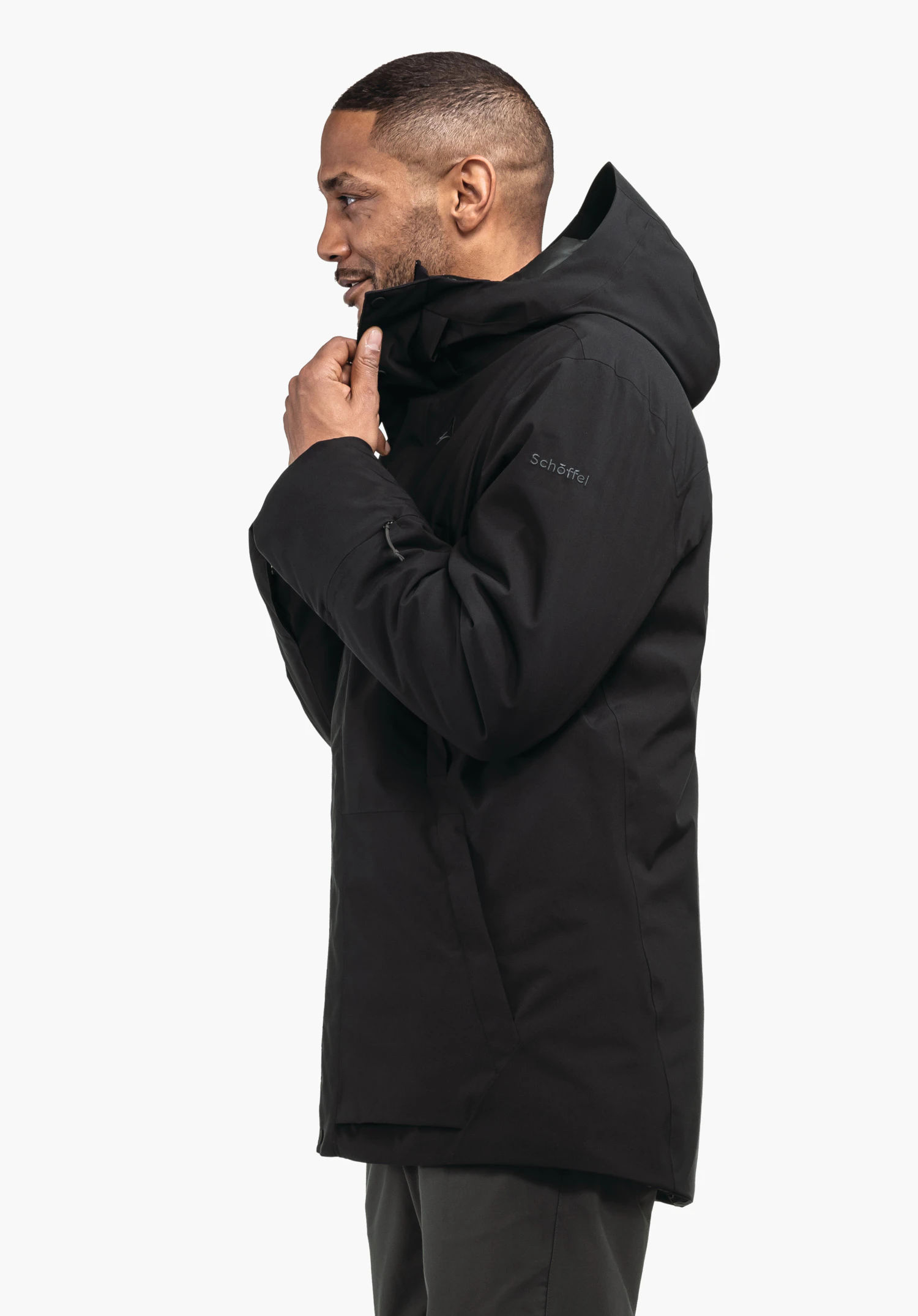 Ins Parka Style Crivat MNS schwarze Parka-Jacke, Mann zieht Kragen hoch; seitliche Aufnahme vor weißem Hintergrund. Text/Logo auf Ärmel: "Schöffel".