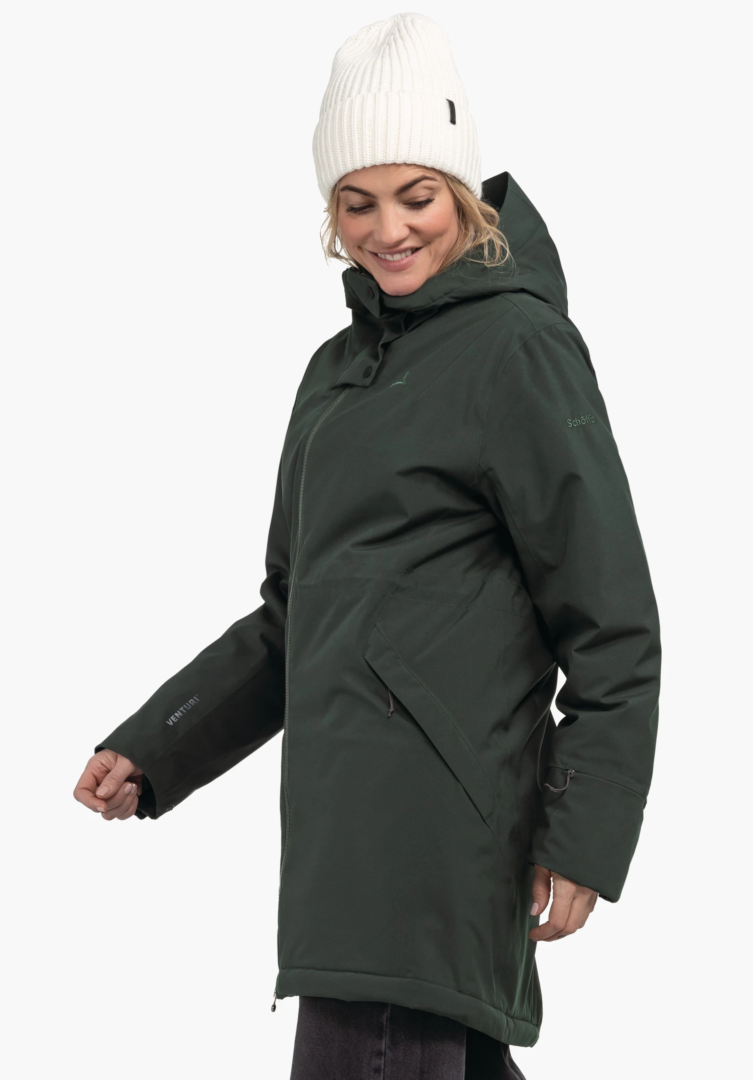 Ins Parka Style Crivat WMS Dunkelgrüner Parka, getragen von einer Frau, die lächelnd nach unten blickt, vor hellem Hintergrund; weiße Mütze. Text auf Ärmel: VENTURI. Text auf Oberarm: Schöffel.
