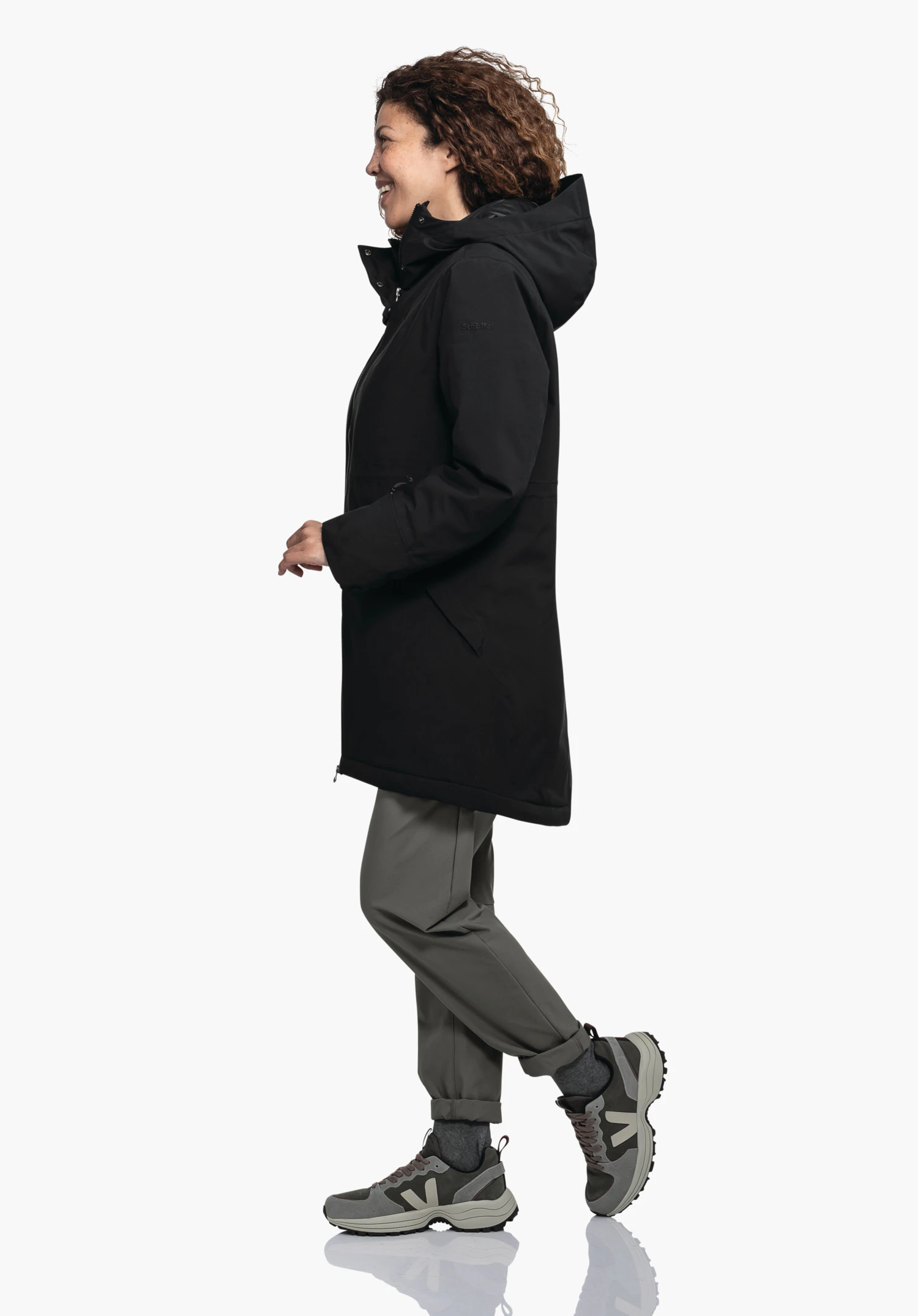 Ins Parka Style Crivat WMS Schwarzer Parka an Frau, die seitwärts nach links geht; lässige Gehpose vor schlichtem weißem Hintergrund.