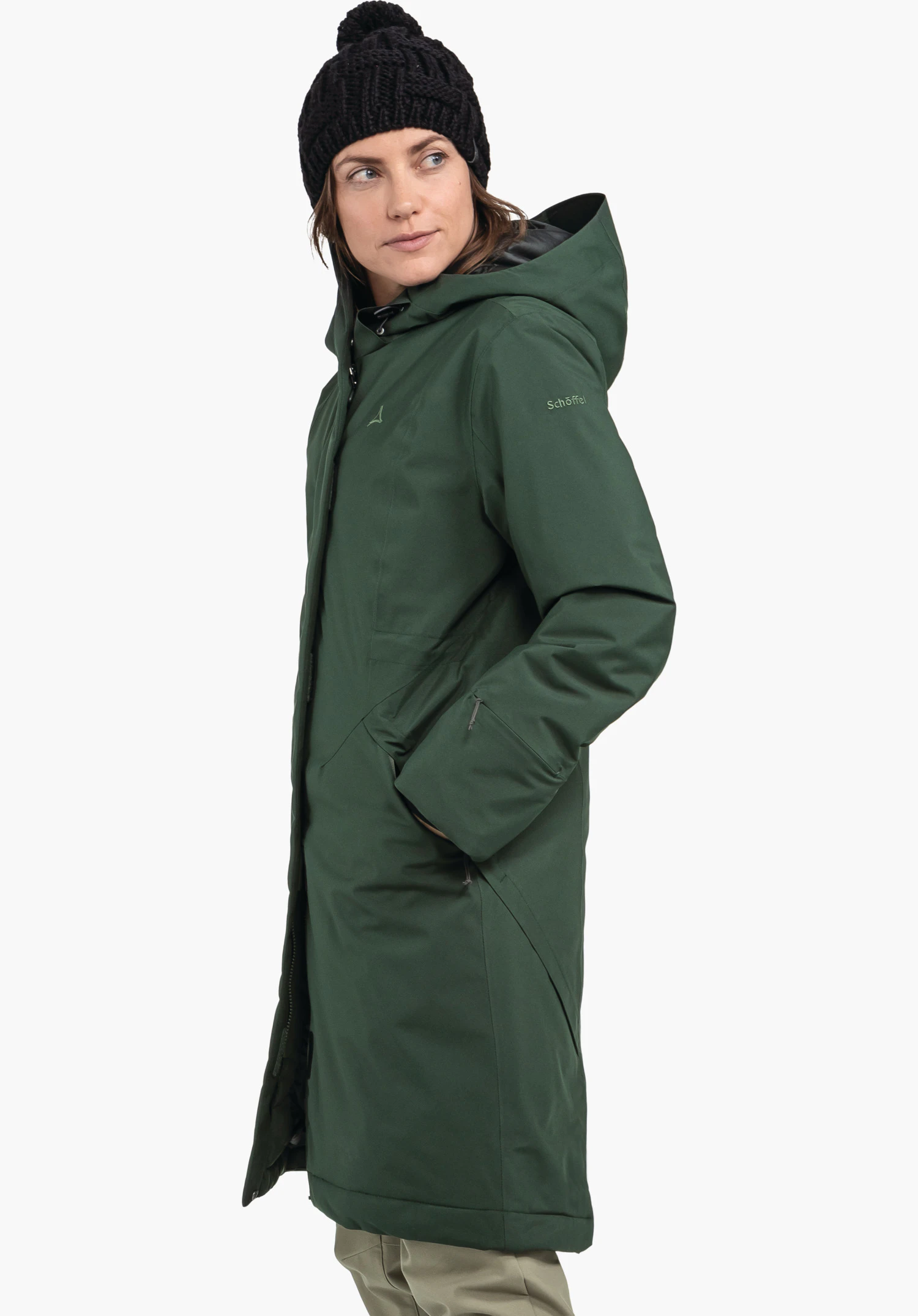 Ins Coat Style Gregale WMS in grün mit Schöffel-Schriftzug, getragen von einem weiblichen Model in der Seitenansicht mit Händen in den Taschen und schwarzer Mütze vor weißem Hintergrund. (KI-generiert)