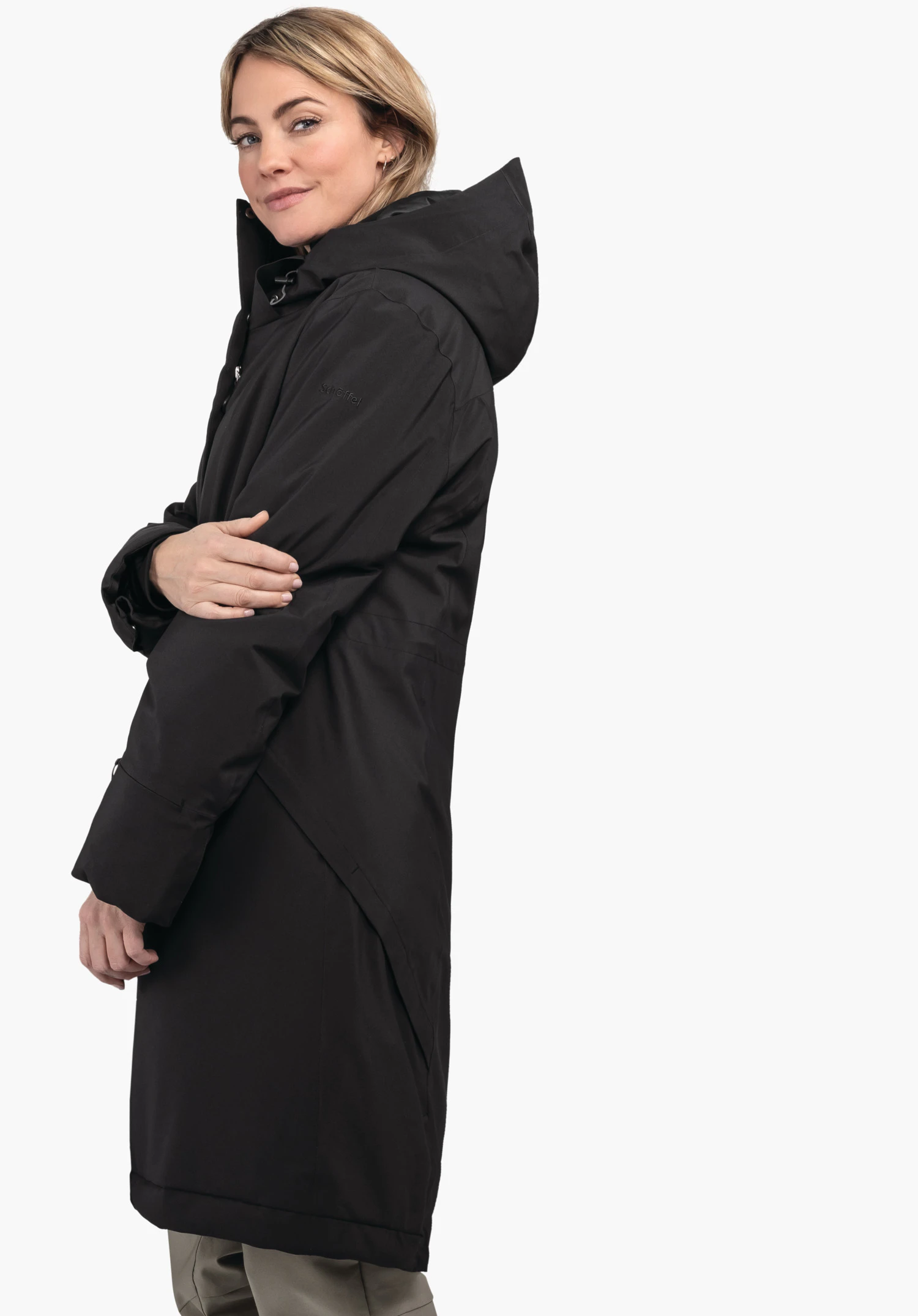 Ins Coat Style Gregale WMS Schwarzer langer Parka mit Kapuze, von einer Person getragen, die die Arme vor der Brust verschränkt und über die Schulter blickt; weißer Hintergrund. Text auf Ärmel: "Schöffel".
