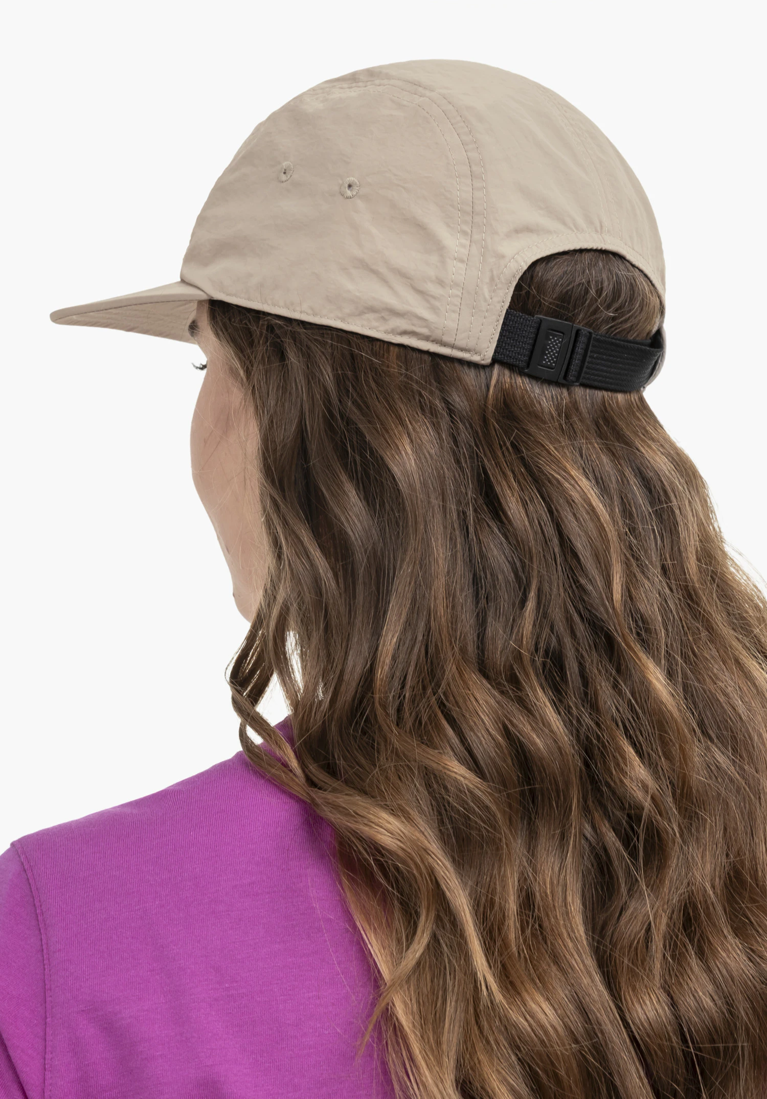 Sun Cap in beige. The model is shown in a half-profile view from behind, highlighting the adjustable closure at the back of the head. Set against a neutral background, the light-coloured cap provides a stylish contrast to the magenta top.