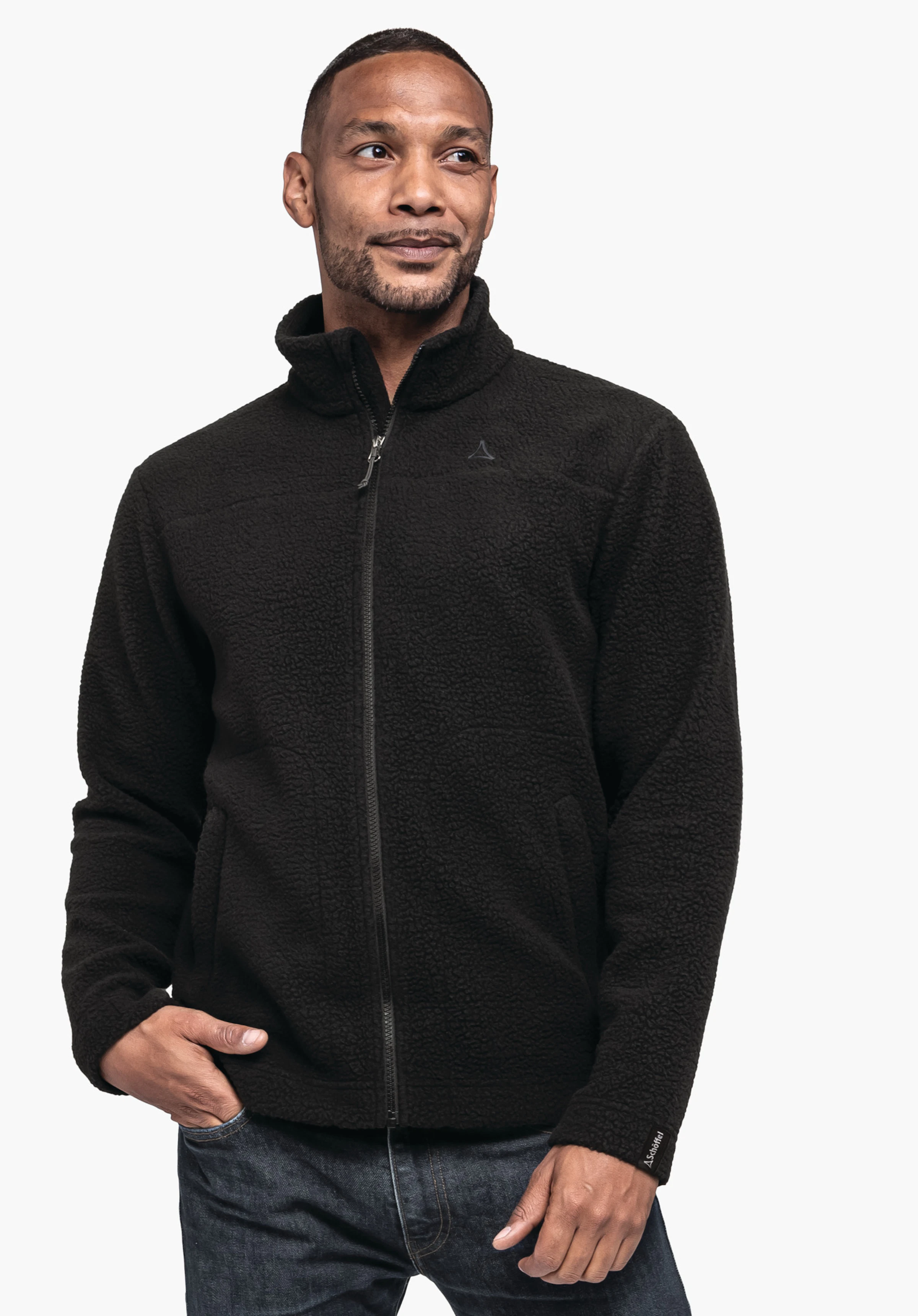 CIRC Fleece Jk Style Purga MNS. Un mannequin porte la veste en polaire de couleur noire avec une fermeture éclair intégrale et le logo Schöffel sur la poitrine gauche. Le mannequin se tient légèrement de profil avec une main dans la poche devant un fond neutre.