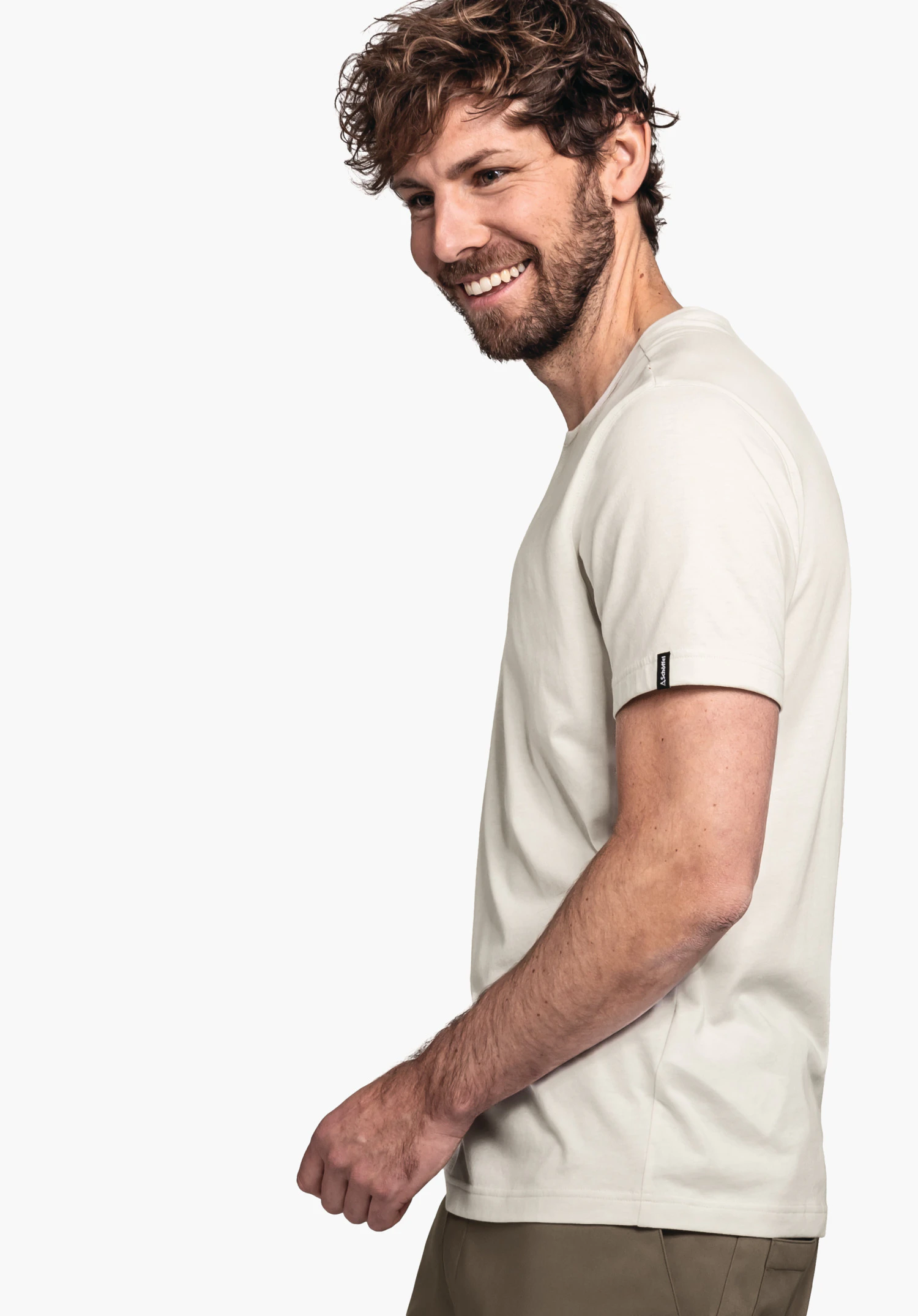 T Shirt Style Collada MNS man met donkerbruin haar glimlacht, staat zijwaarts; draagt een lichtwit T-shirt en bruine broek voor een lichte neutrale achtergrond. Geen tekst.