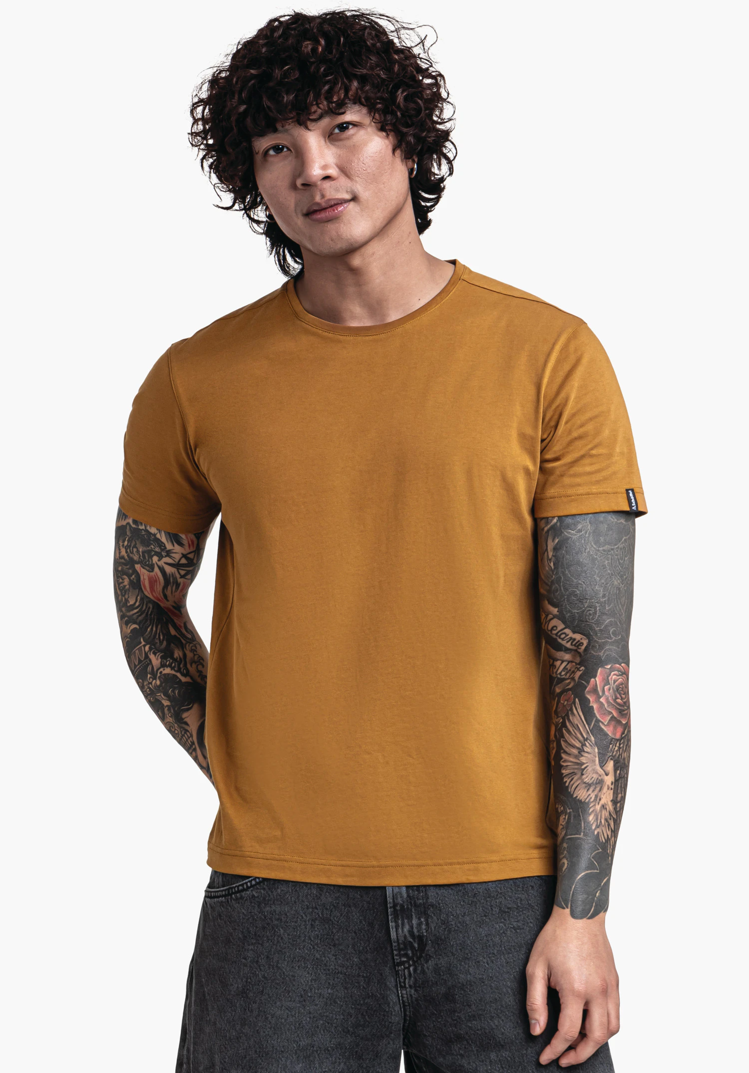T Shirt Style Collada MNS T-shirt orange foncé à manches courtes porté par un homme debout aux bras tatoués, regard légèrement de côté; fond clair neutre.