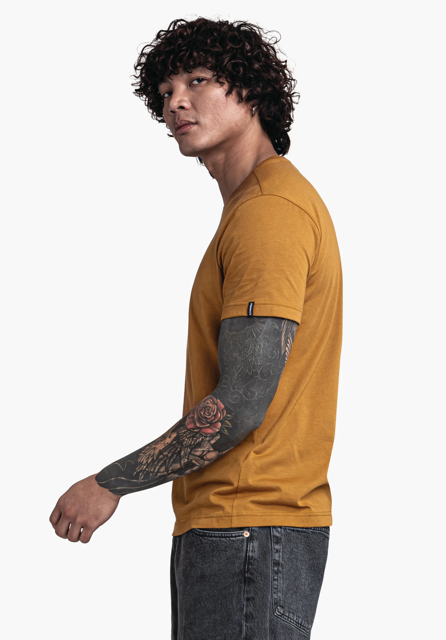 T Shirt Style Collada MNS t-shirt jaune foncé porté par un homme; il se tourne de profil, regard vers la caméra, bras tendu avec tatouage noir et rose rouge; fond clair neutre.