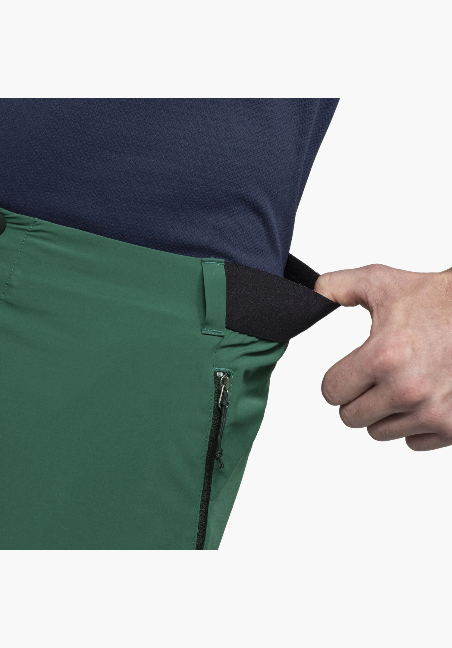 Pants Style Blaustein MNS, avec une main démontrant l'empiècement extensible noir sur la ceinture verte, à côté d'une poche latérale zippée, gros plan sur fond blanc/neutre.