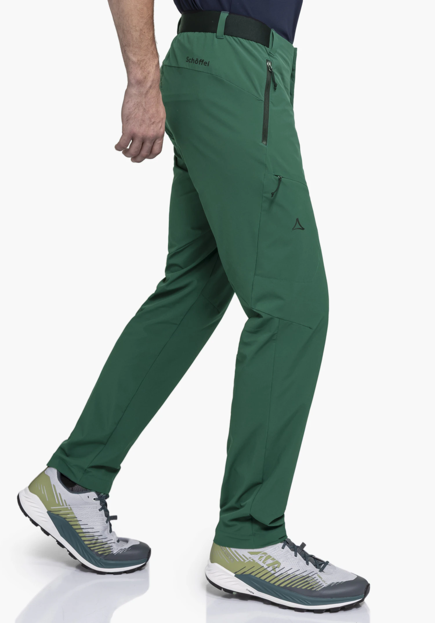 Pants Style Blaustein MNS en vert. Le mannequin se tient de profil dans une pose de marche. Une poche latérale zippée et le logo Schöffel sur la cuisse droite et à l'arrière de la taille sont visibles devant un fond neutre.
