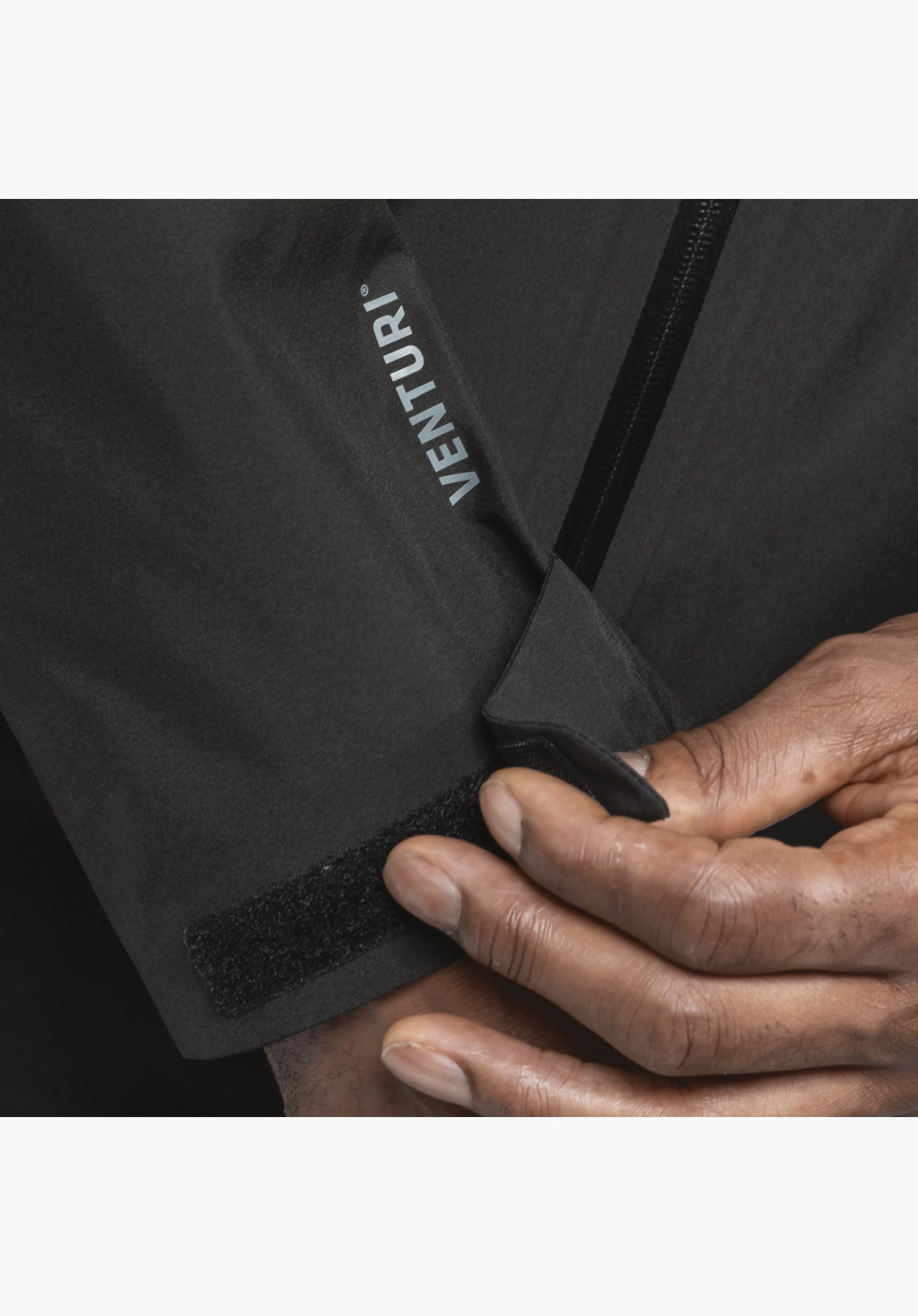Jacket Style Mauku MNS, detail of the adjustable hook-and-loop fastener on the black sleeve cuff being held by a hand, featuring a visible VENTURI print on the fabric, close-up against a white/neutral background.