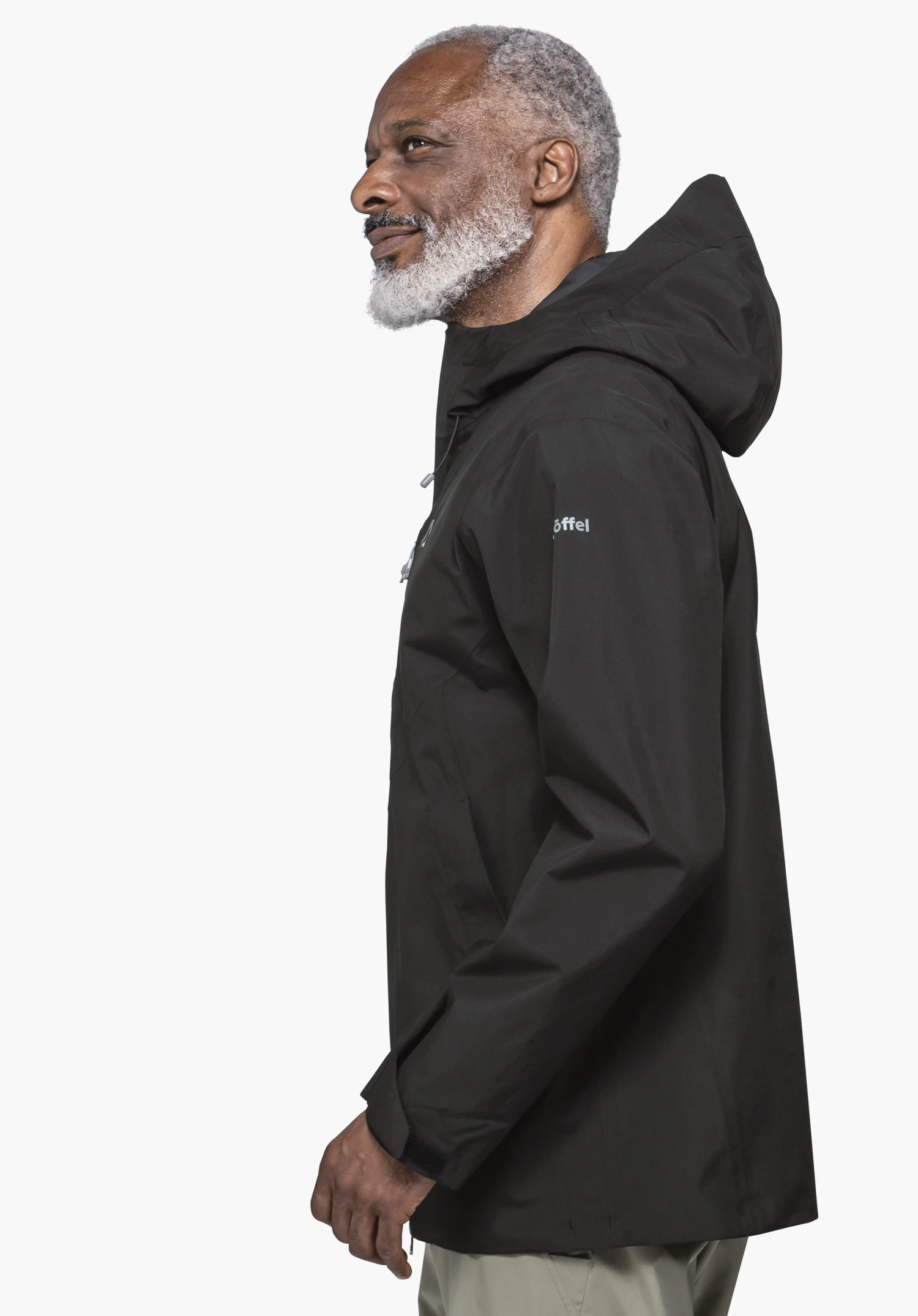 Jacket Style Mauku MNS in black captured against a neutral background. The model is standing in a side profile, showcasing the functional jacket and its hood. The Schöffel-logo on the upper arm is clearly visible.