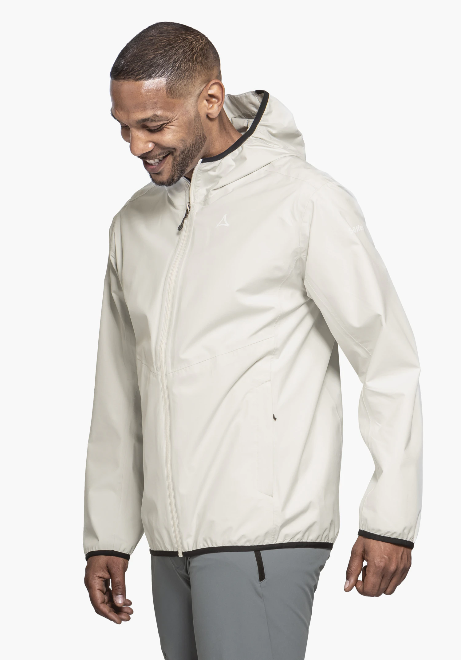 Jacket Style Migandi MNS getragen von einem lächelnden Model, das leicht seitlich steht und nach unten blickt. Die geschlossene Jacke in der Farbe Natur zeigt das dezente Schöffel-Logo auf der linken Brust, präsentiert vor einem neutralen Hintergrund.