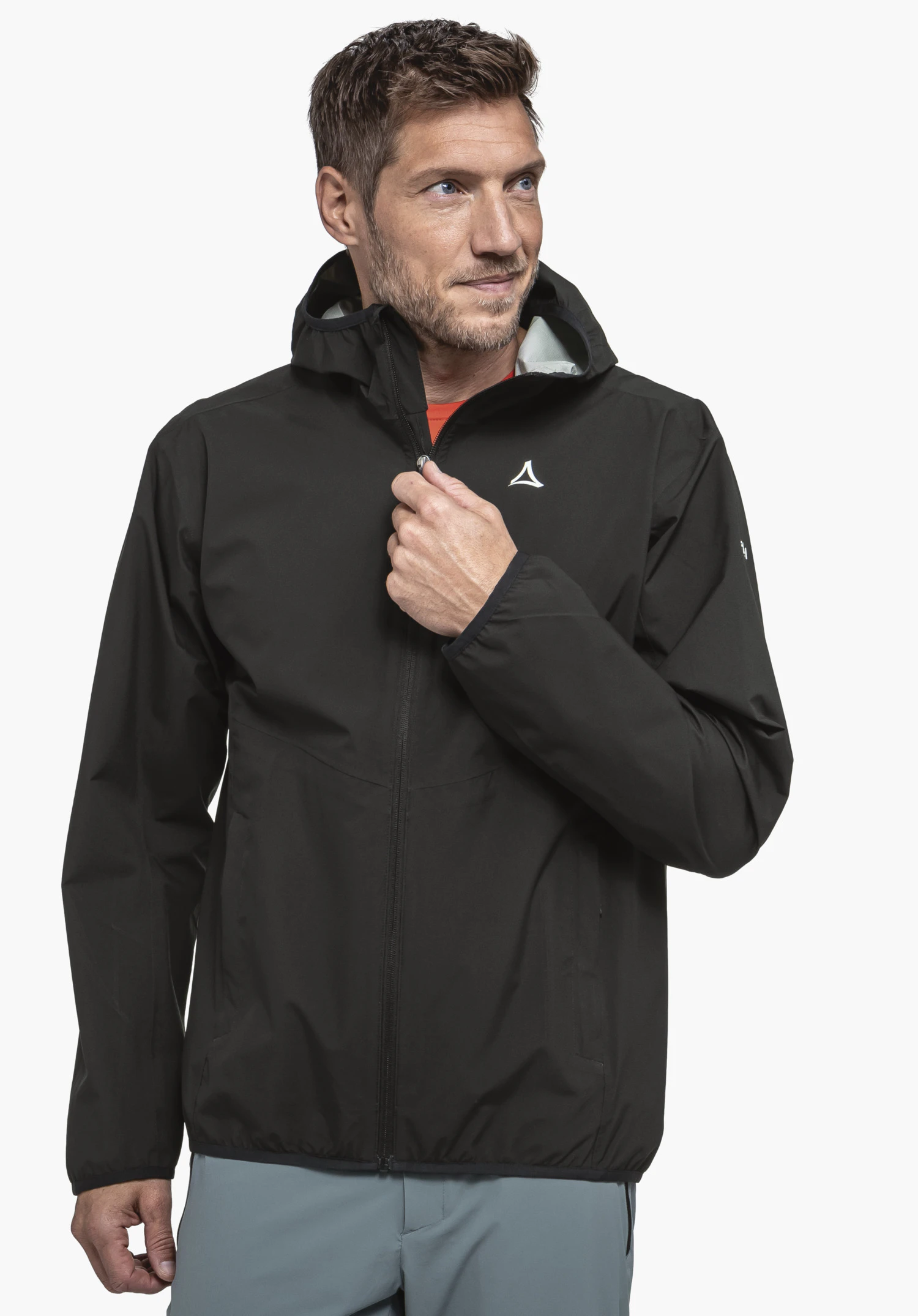 Jacket Style Migandi MNS Veste de pluie noire avec logo Schöffel, homme remontant le zip; t-shirt rouge visible, doublure de capuche blanche, pantalon bleu clair, fond blanc, photo studio.