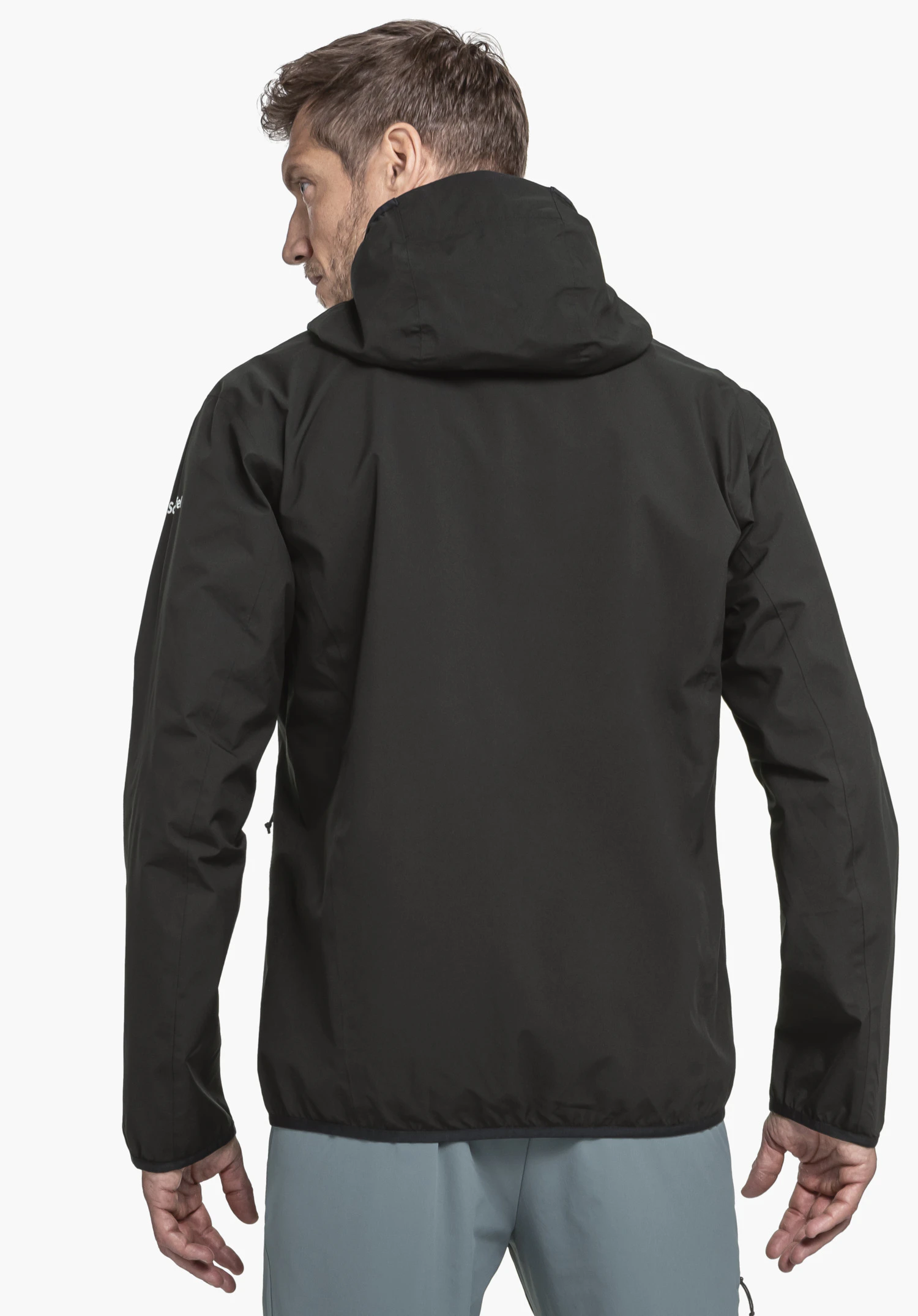 Jacket Style Migandi MNS black rain jacket worn by a man from the back; hood up, light blue trousers; neutral white studio background.