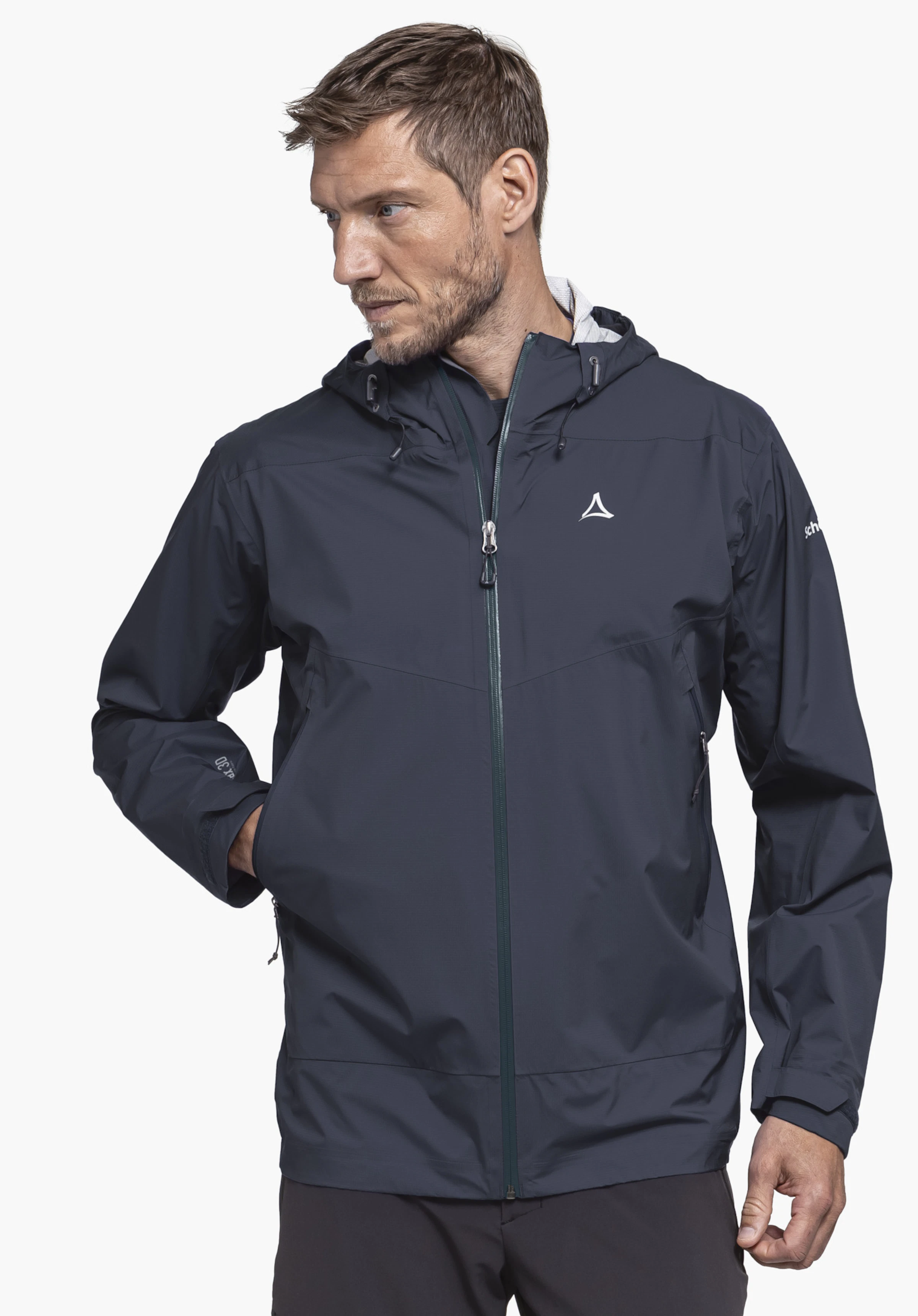 2.5L 3D Jacket Vistdal M getragen von einem Model, das zur Seite blickt und eine Hand in der Tasche der blauen Jacke hat. Auf der Brust ist das Schöffel-Logo zu sehen, fotografiert vor einem neutralen Hintergrund.