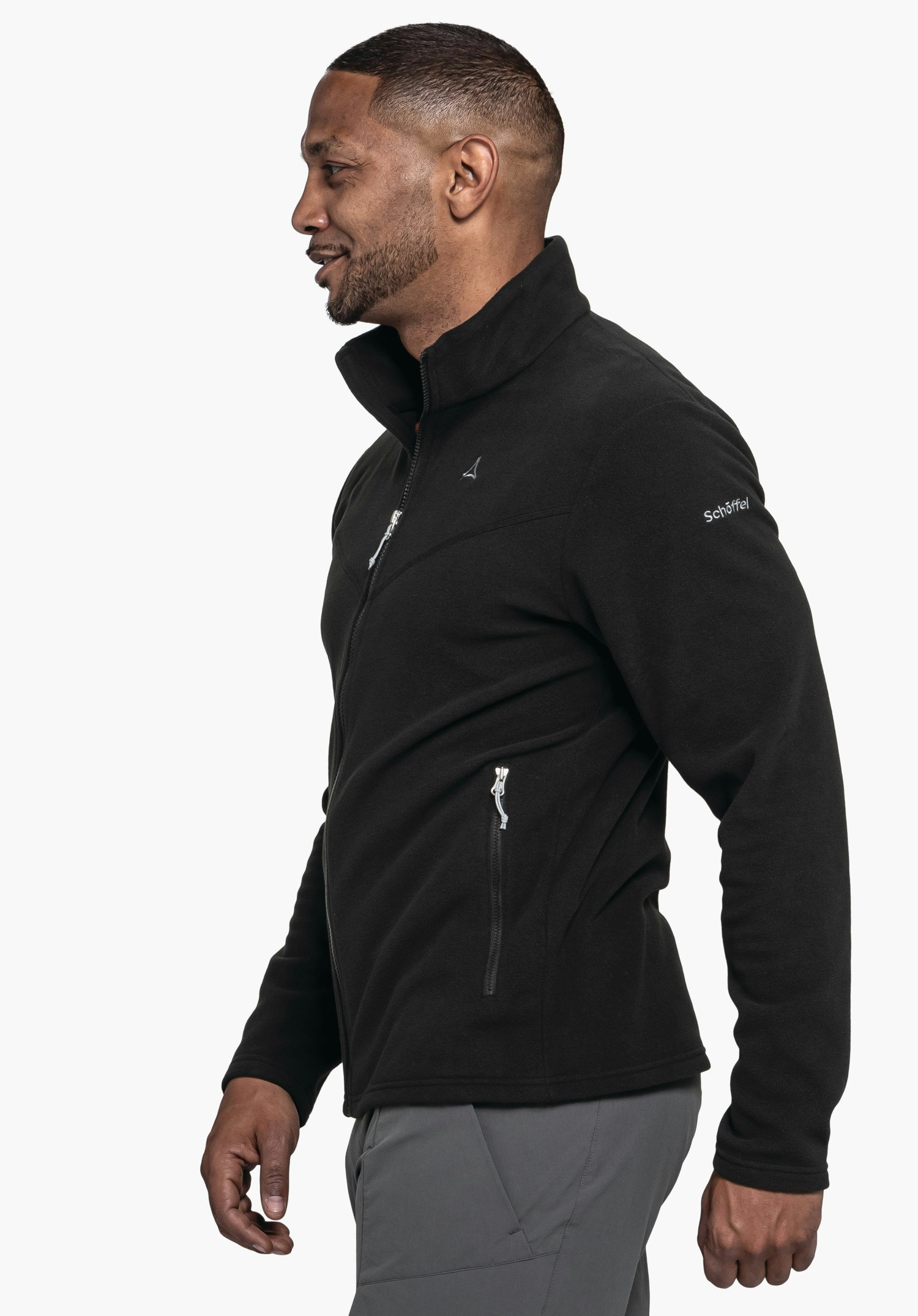 Fleece Jk Style Qutang MNS Schwarze Fleecejacke mit Schöffel-Logo auf der Brust, getragen von einem Mann im Profil, steht leicht lächelnd; Reißverschlüsse sichtbar, Ärmelbeschriftung: "Schöffel". Kontext: neutraler weißer Hintergrund, dunkelgraue Hose.