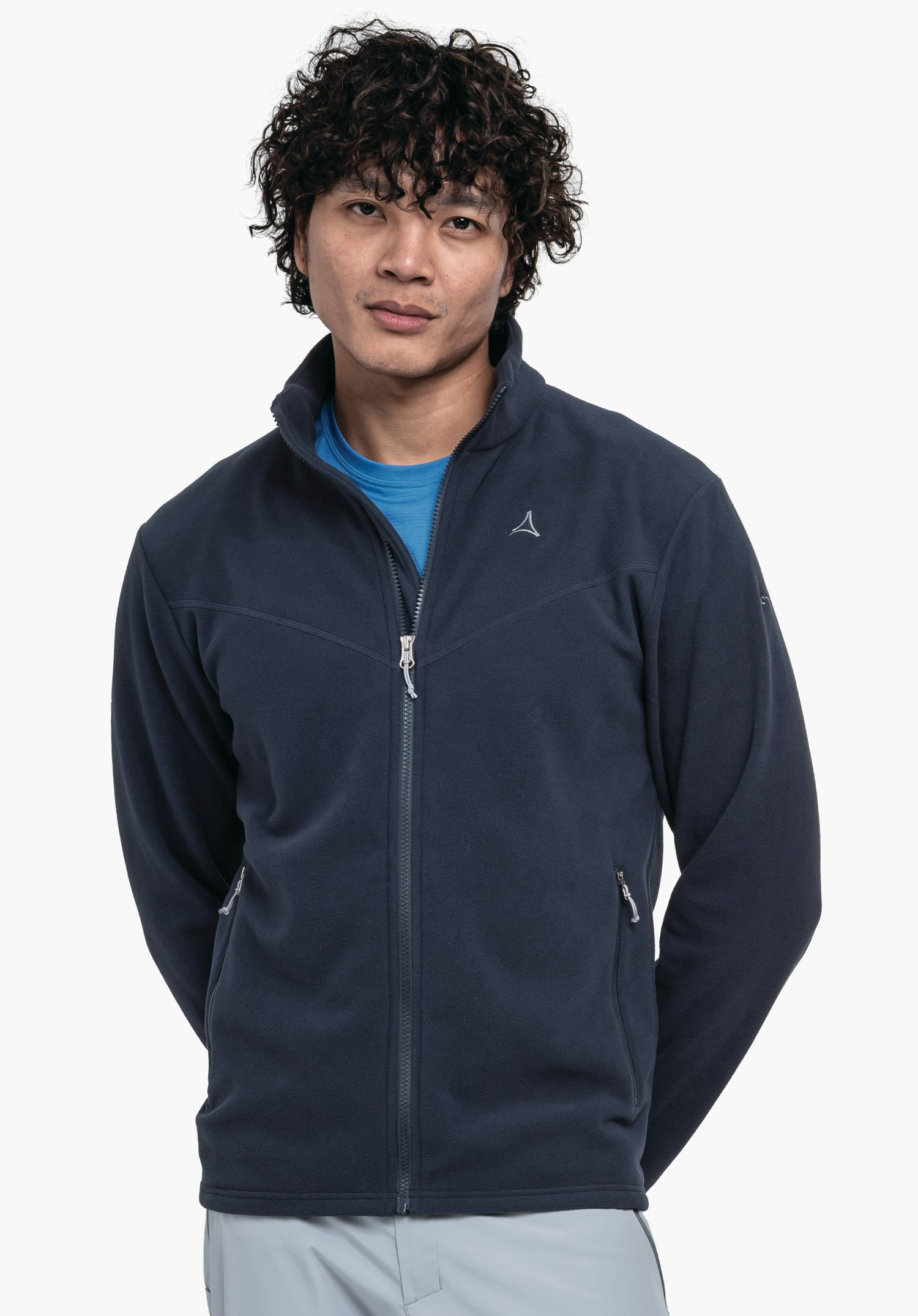 Fleece Jk Style Qutang MNS dunkel blaue Fleecejacke mit Schöffel-Logo, getragen von einer Person, steht mit den Händen hinter dem Rücken, leicht lächelnd vor weißem Hintergrund; hell blaues Shirt und hell graue Hose sichtbar.