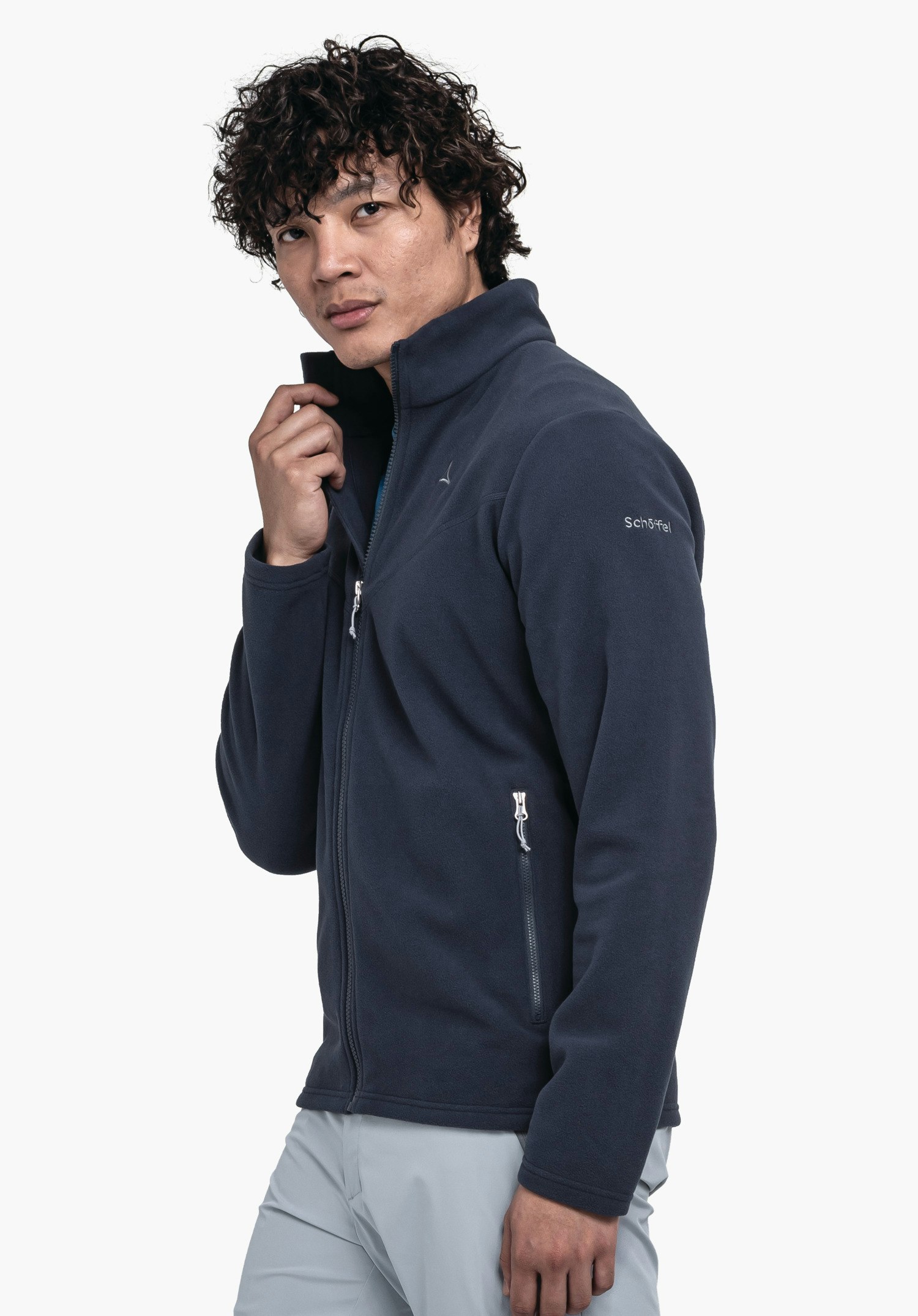 Fleece Jk Style Qutang MNS Mann trägt Fleecejacke in dunkel blau, hält Kragen mit rechter Hand; steht vor weißem Hintergrund. Hose in hell grau. Text auf Ärmel: "Schöffel".