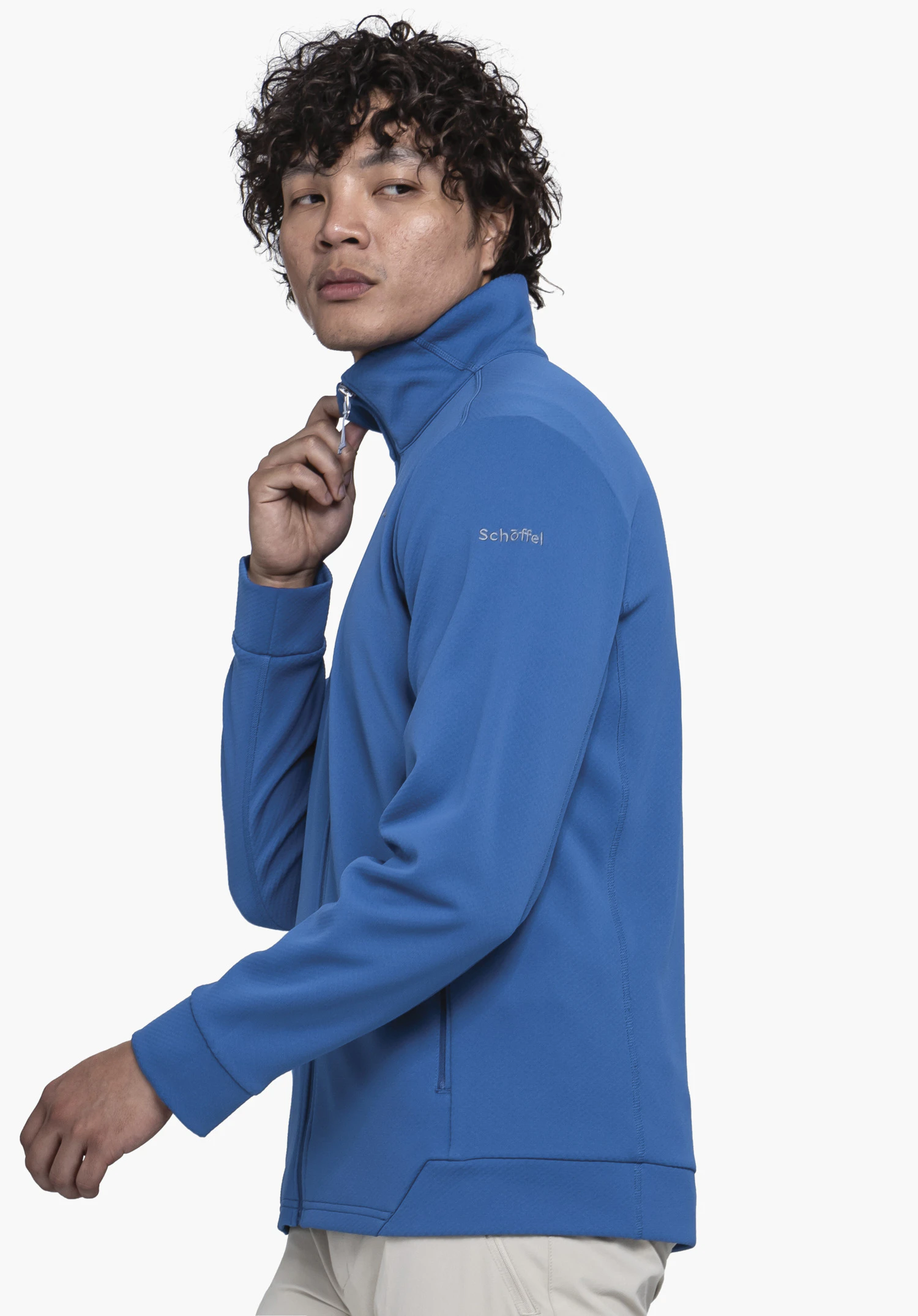 Fleece Jk Style Uriezzo MNS en bleu. Un modèle se tient de profil sur un fond neutre et remonte la fermeture éclair de la veste en polaire, le logo Schöffel étant bien visible sur le haut du bras.